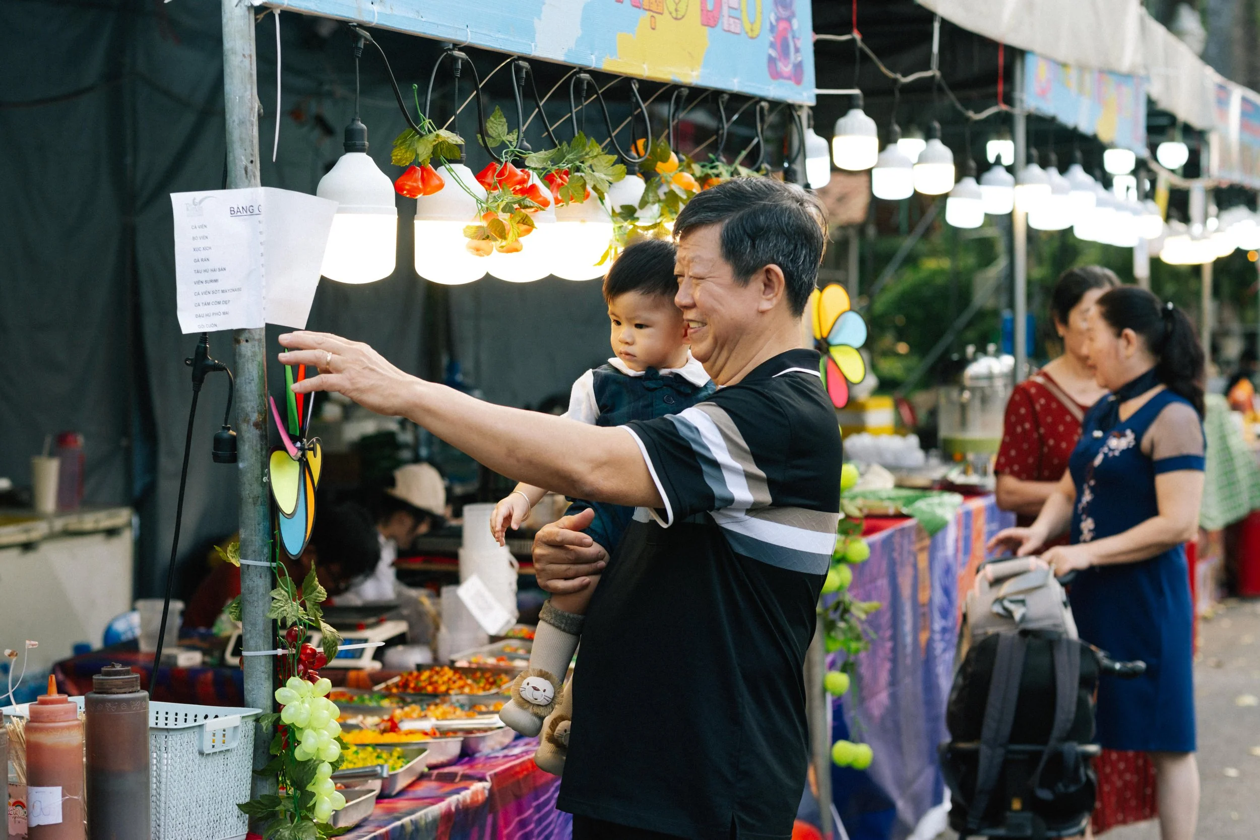 A smiling man holding a young child while shopping at an outdoor market, with women browsing stalls in the background.