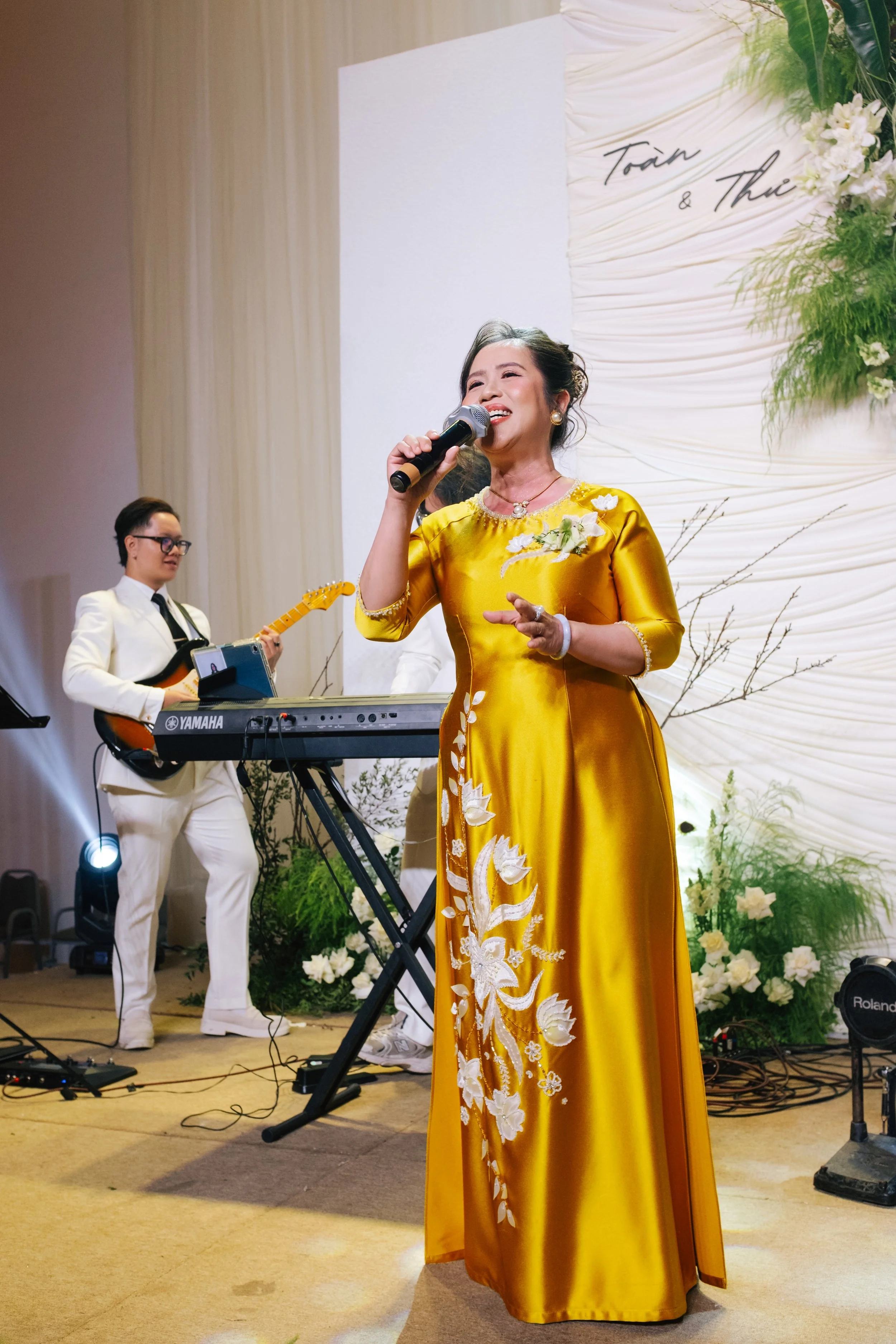 A woman in a yellow traditional dress singing into a microphone on stage, accompanied by a musician playing keyboard and guitar at a wedding or celebration event.