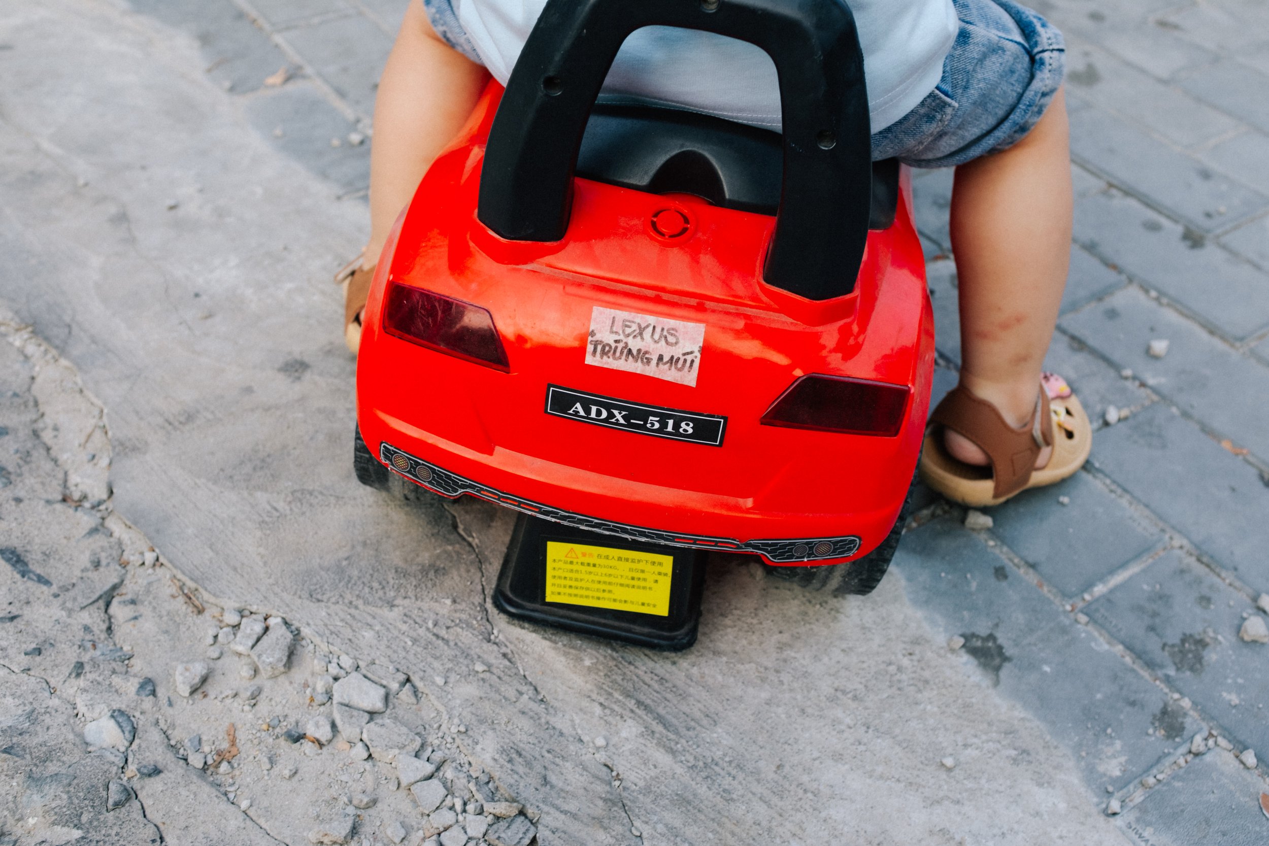 Children playing with a red toy car on a sidewalk, visible only from the back, with one child's legs and sandals showing.