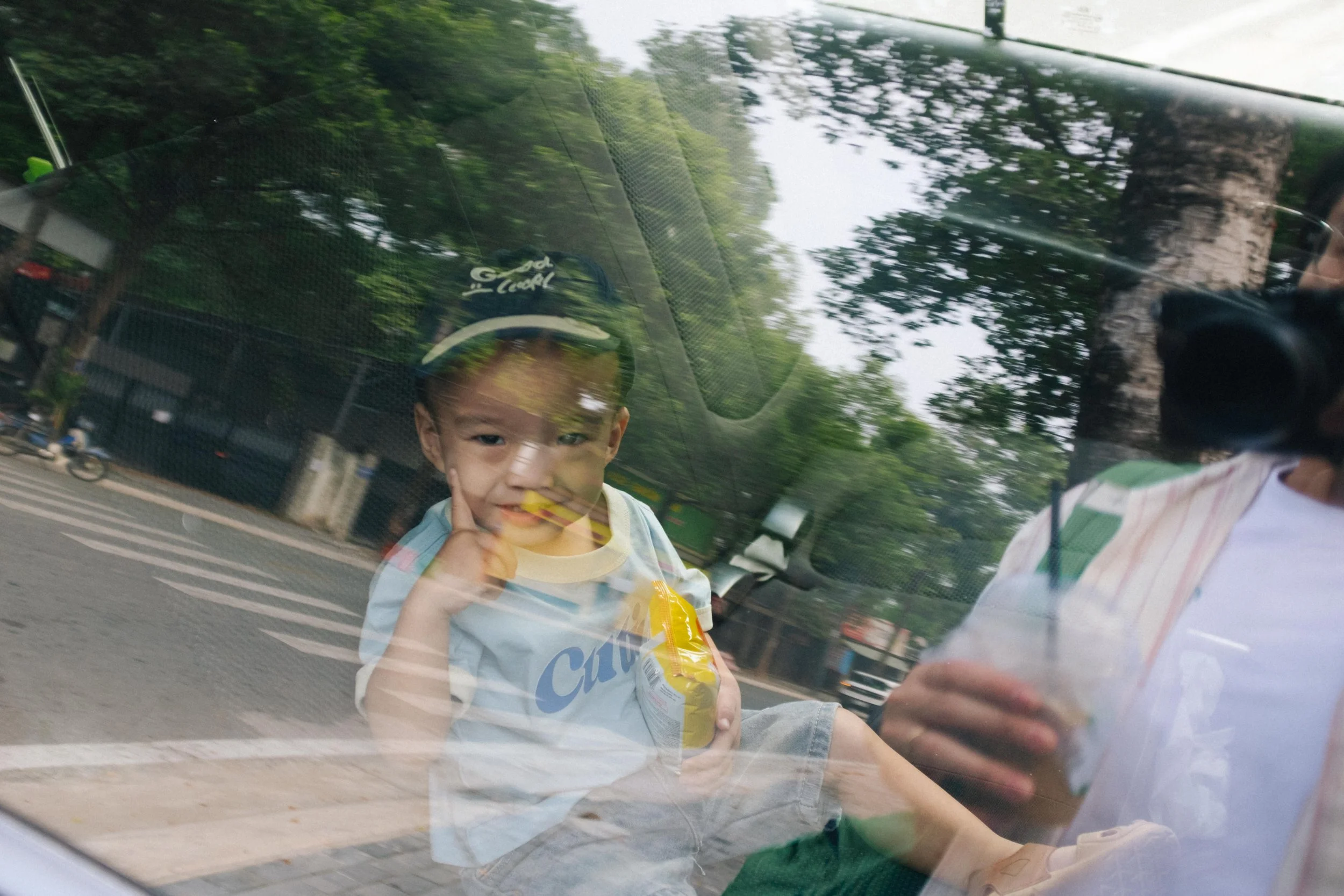 Reflection of a young boy smiling and resting his finger on his cheek while sitting inside a vehicle, with outdoor trees and street scene visible through the window.