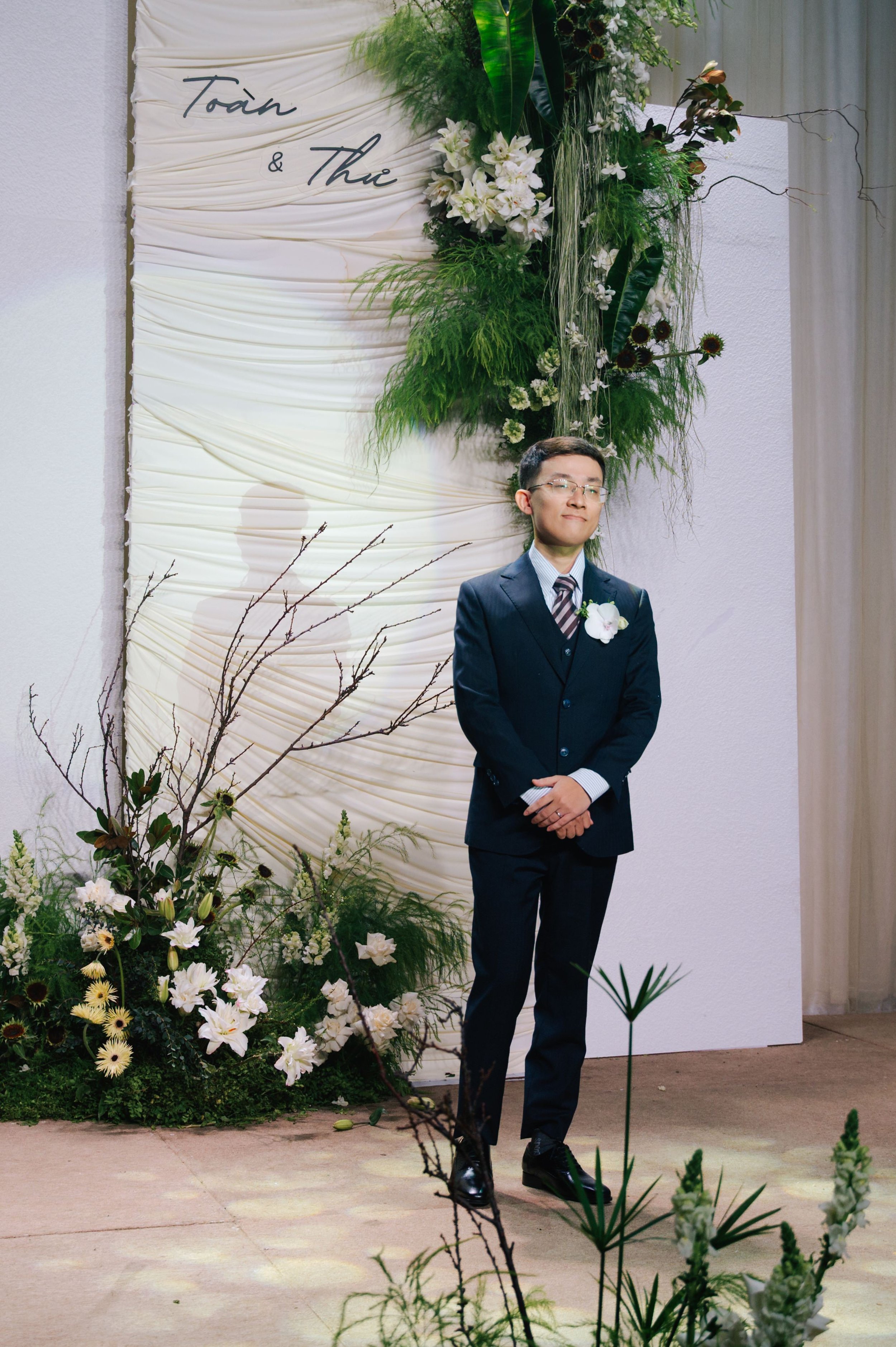 A man in a dark suit, white shirt, and striped tie standing with hands clasped in front, at a wedding ceremony with floral decorations and a white fabric backdrop that has handwriting text.