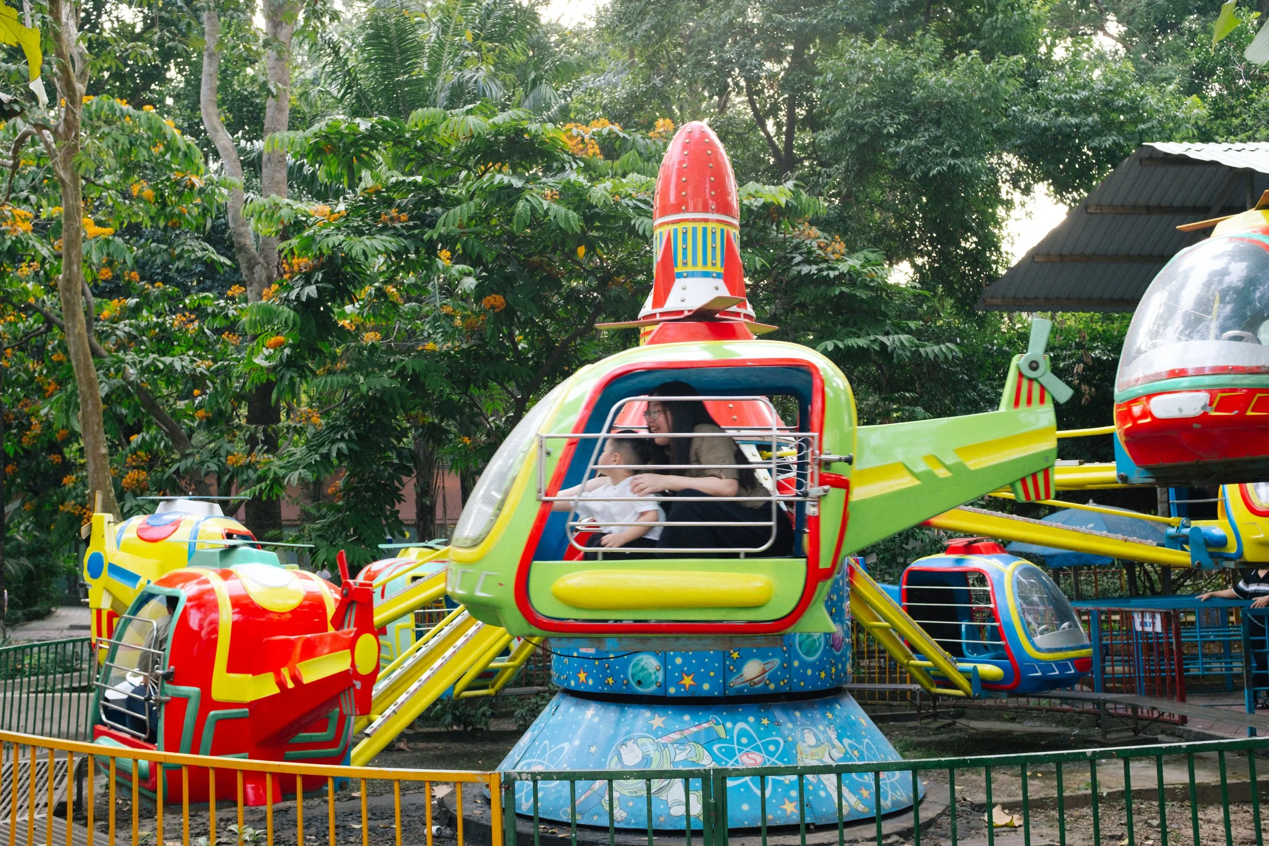 Children and a woman riding a space-themed amusement park ride shaped like a spaceship, surrounded by green trees.