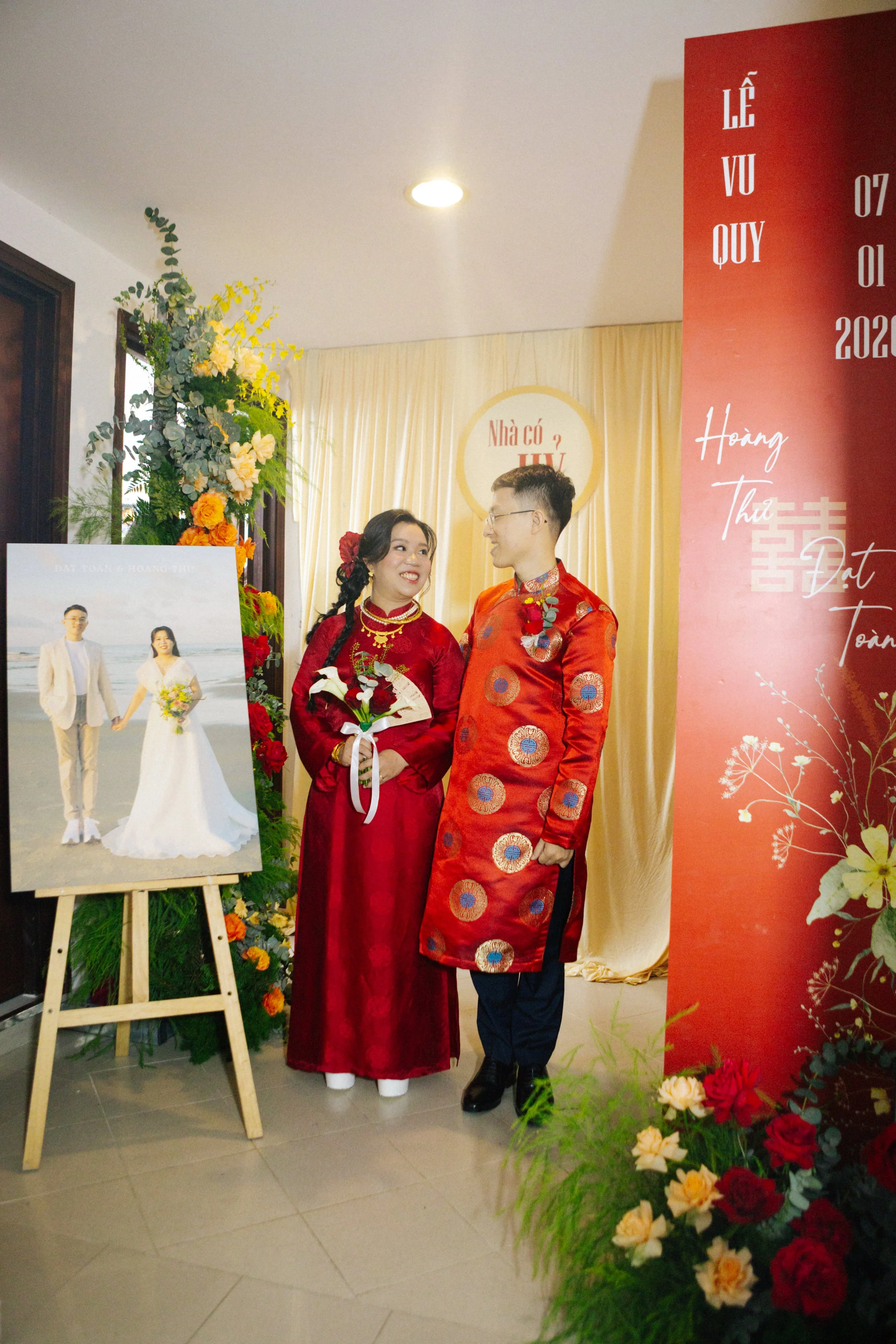 A couple in traditional Vietnamese wedding attire standing indoors near wedding decor, with a large wedding photograph on an easel and floral arrangements around them.