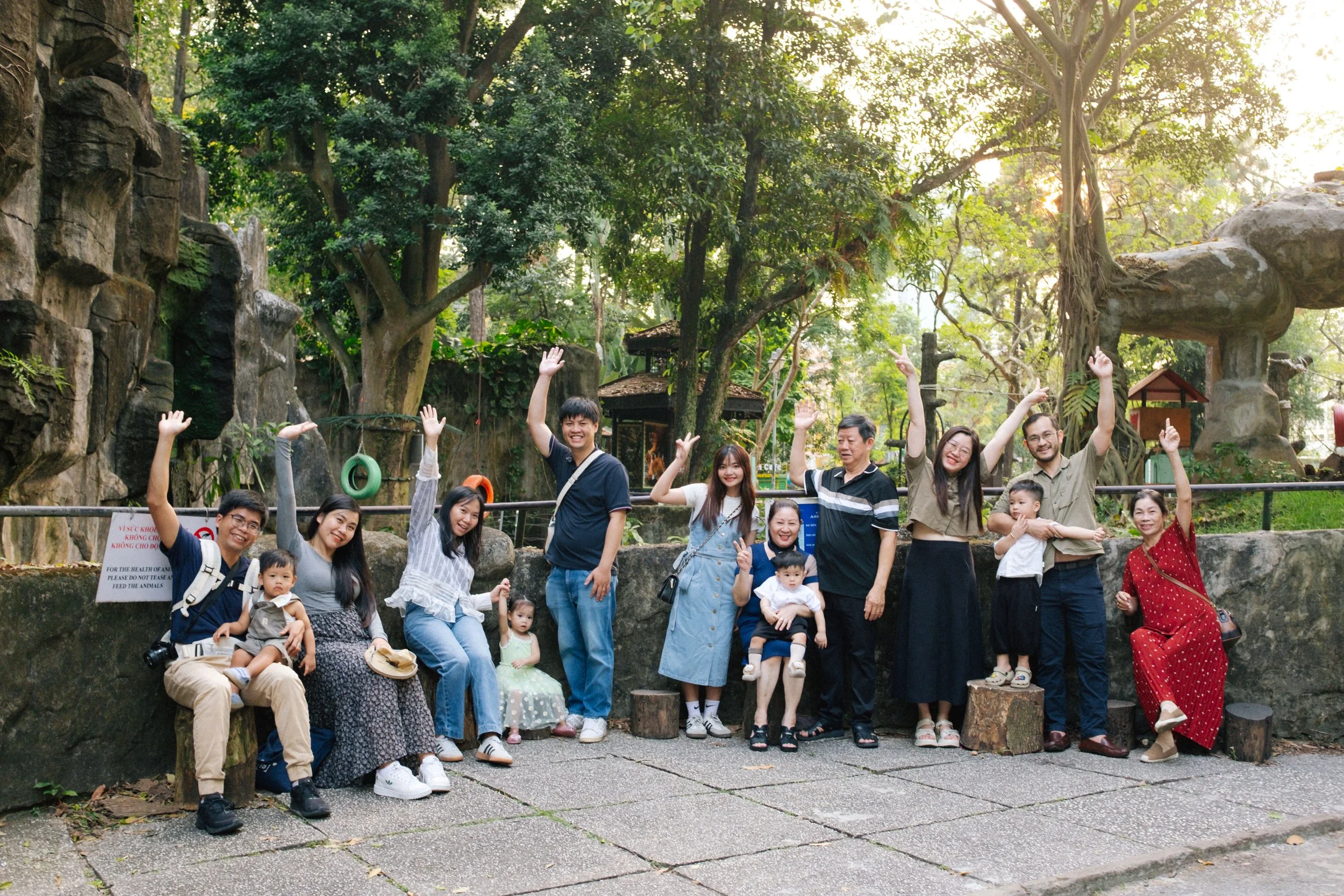 A group of 12 people, including children, smiling and waving at the camera, standing outdoors in a lush, green park with rocks and trees in the background.
