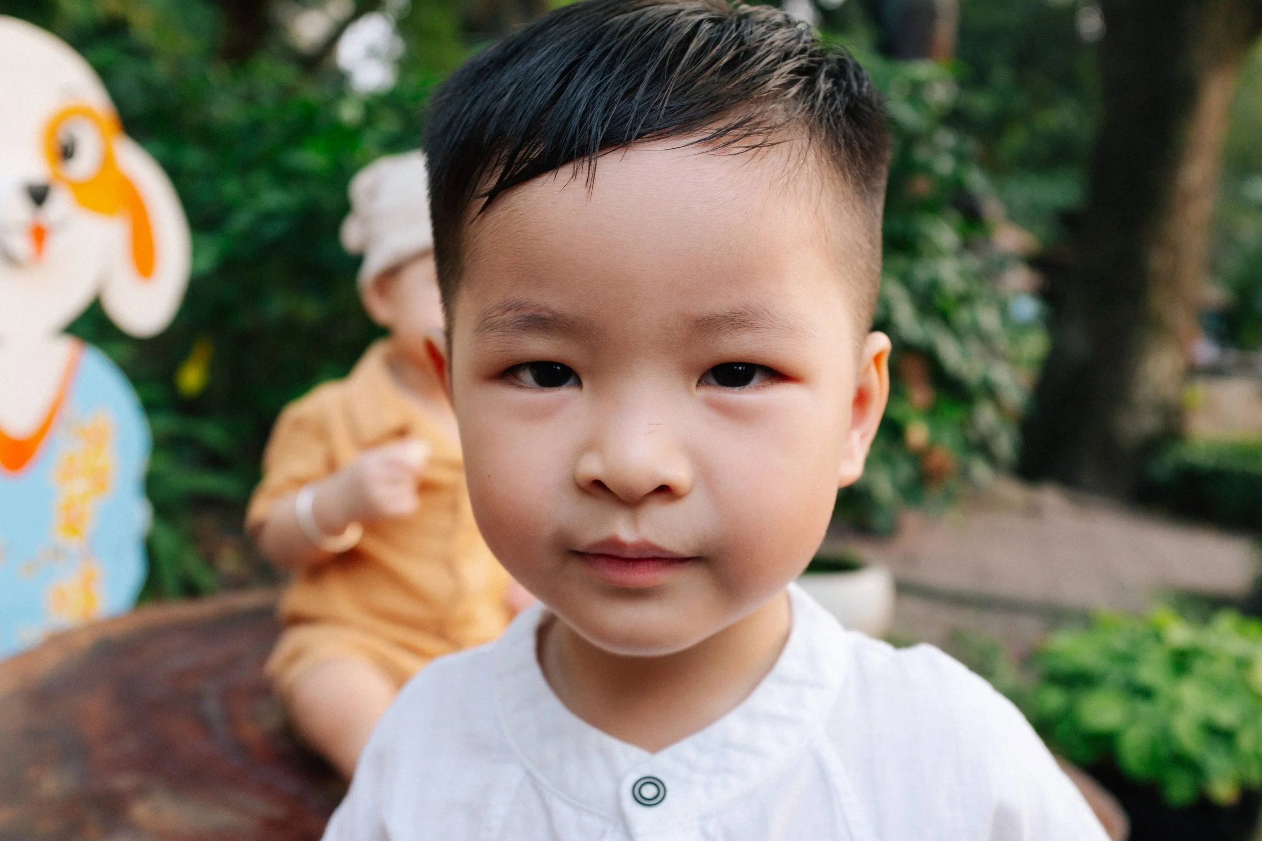 Close-up of a young Asian boy with short black hair, wearing a white shirt, outdoors in a garden or park.