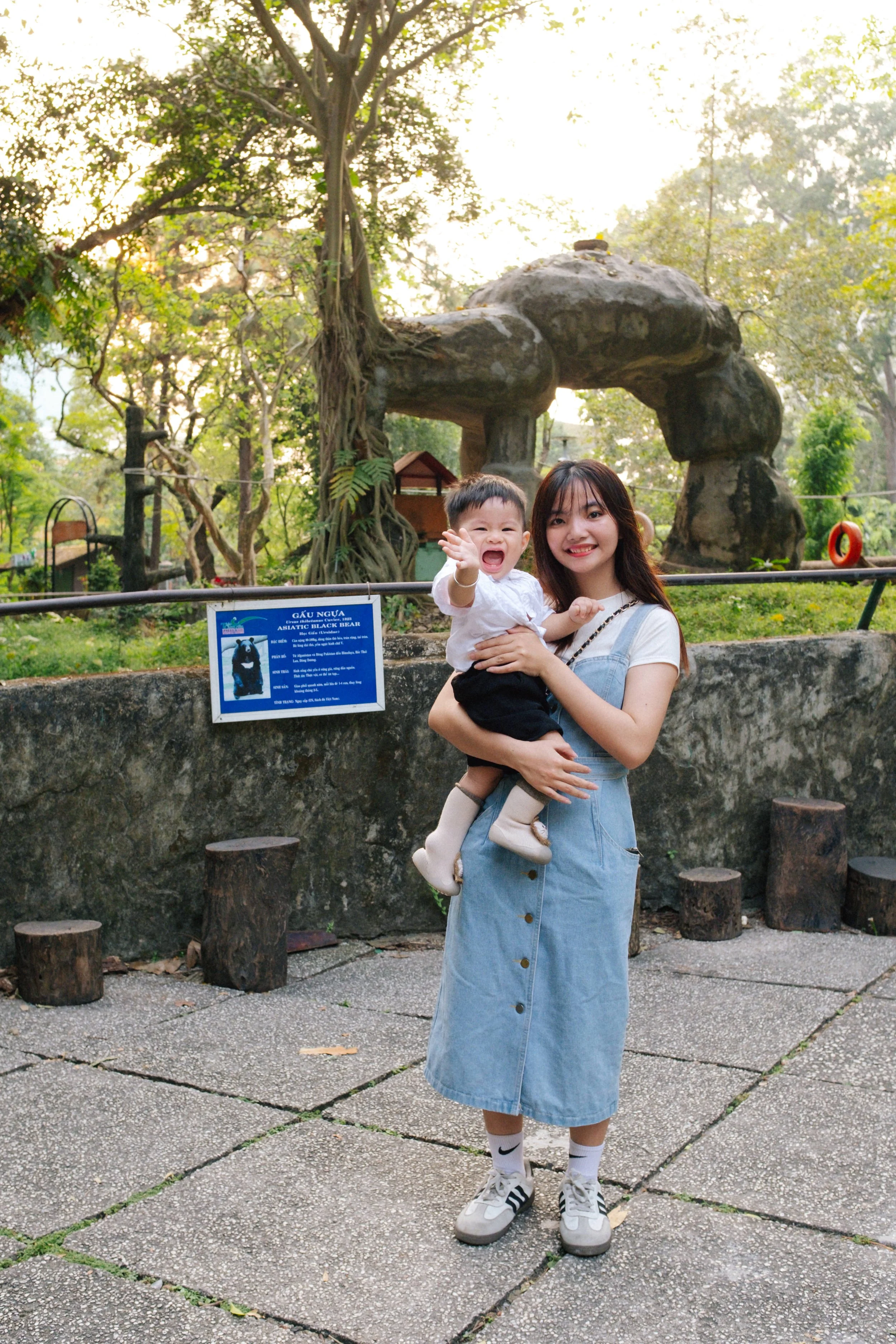 A woman holding a crying child outdoors in front of a zoo exhibit with a sign about a black bear and a rock formation. The woman is smiling, wearing a white shirt, denim dress, and sneakers. The child is wearing a white shirt and black shorts.