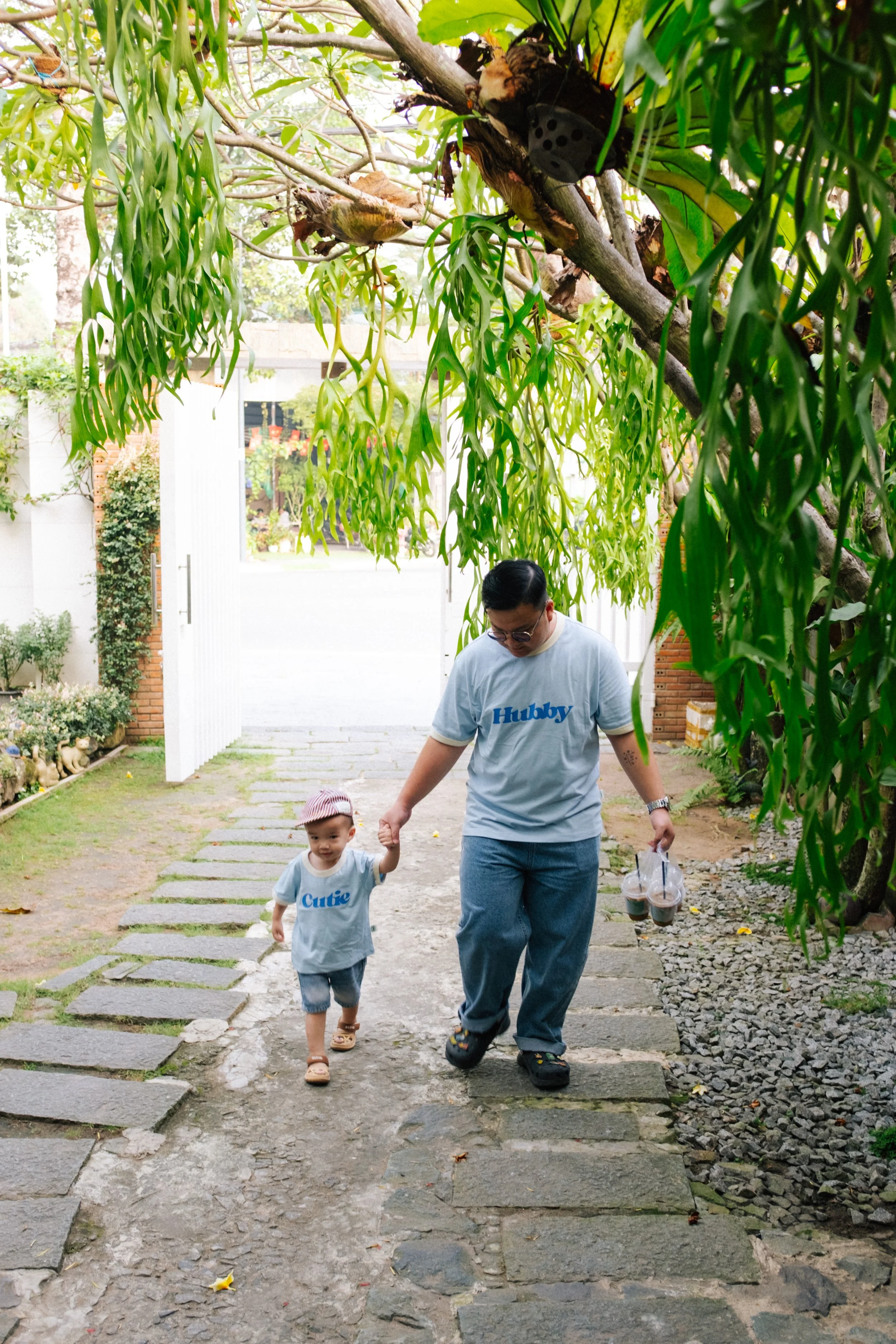 A man and a young child walking hand in hand outdoors on a stone path under a large tree with hanging green leaves. The man is holding drinks, and they are both wearing matching shirts that say 'Hubby' and 'Cutie'.