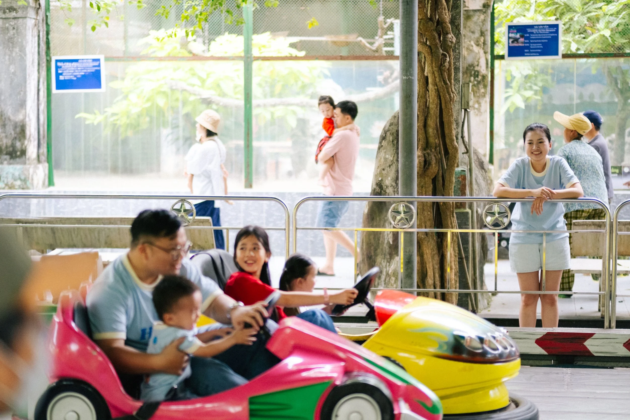 People enjoying a bumper car ride at an amusement park, with children and adults sitting in colorful cars, and others standing or walking nearby.