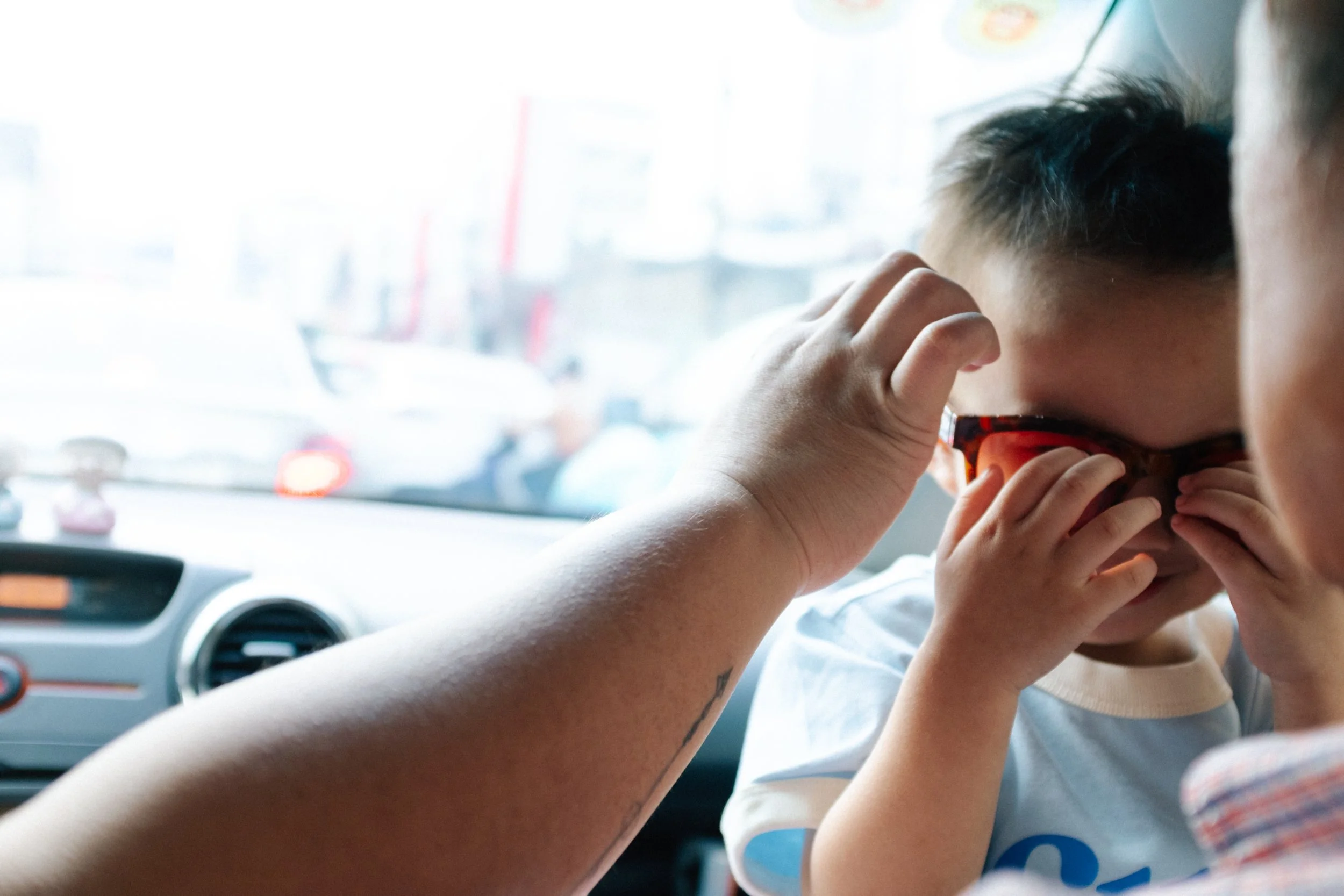 A young boy is adjusting his sunglasses while sitting in a vehicle, with an adult close to him. The background shows a blurry view of traffic outside the vehicle.