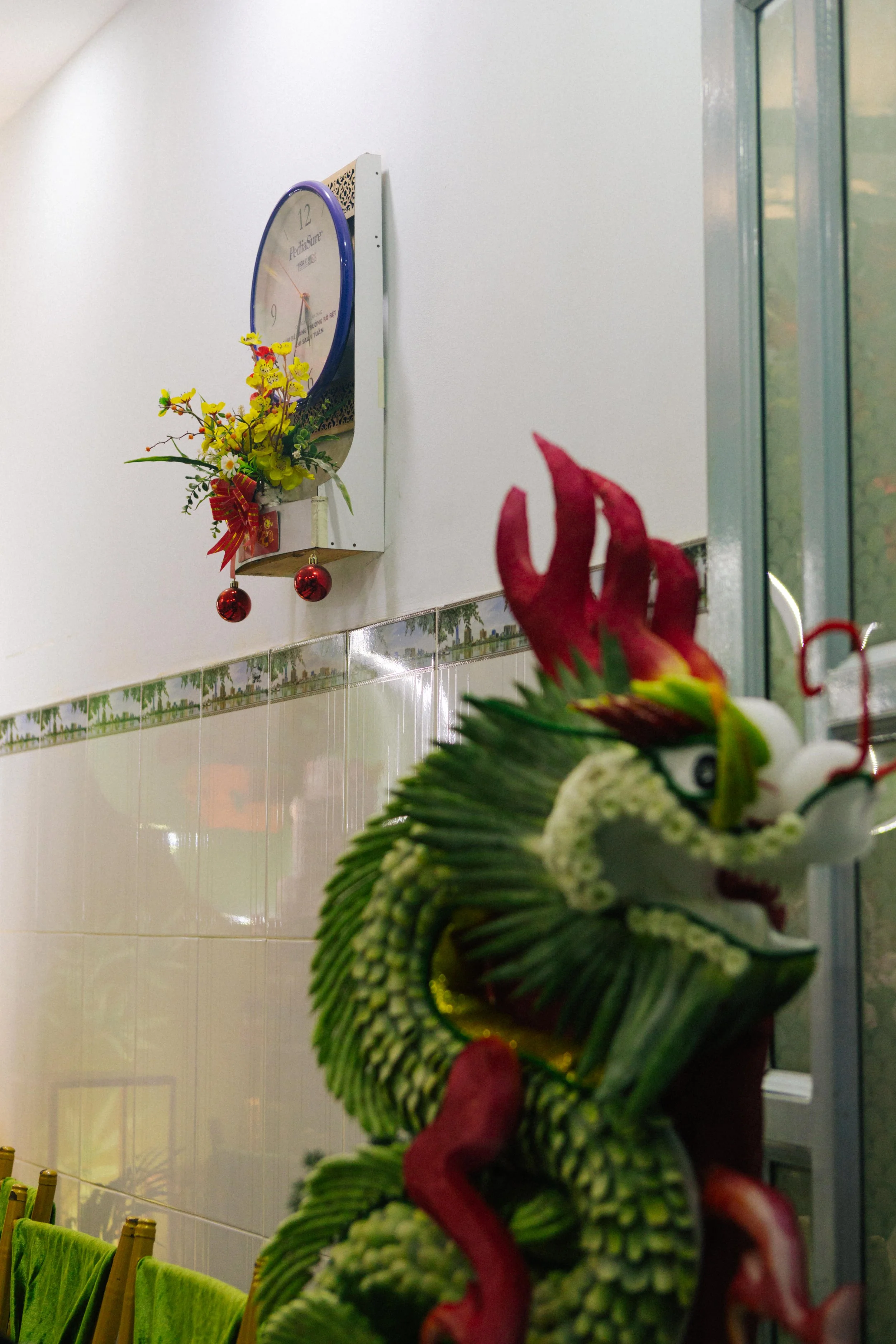 Close-up of a decorative ceramic dragon sculpture with a detailed face, green scales, red horns, and a fierce expression, positioned near a glass door in a room with a wall clock and a flower arrangement visible in the background.