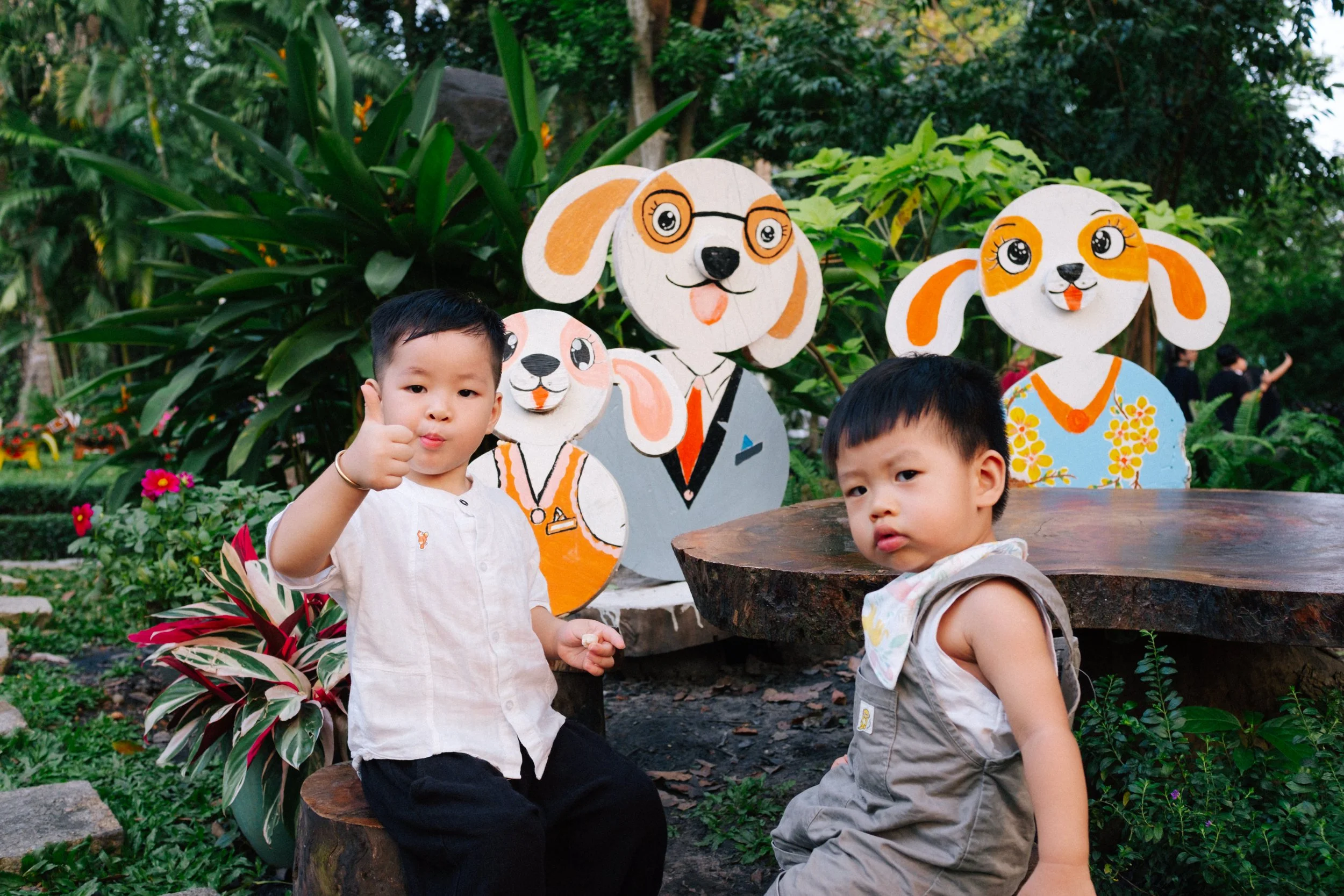 Two young boys sitting outdoors in front of colorful cartoon dog and puppy cutouts, with lush green plants and flowers in the background.