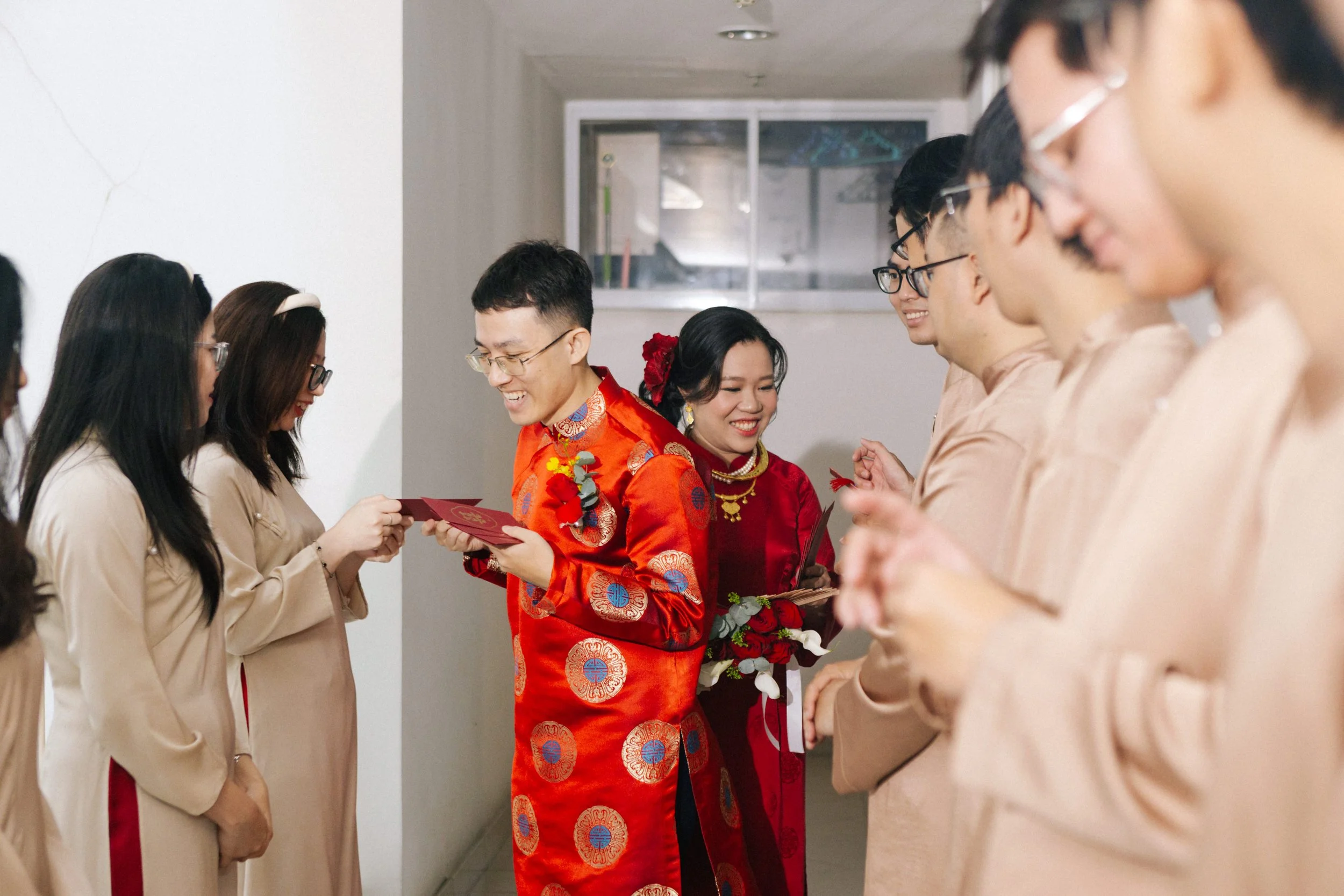 A traditional Chinese wedding ceremony with a bride and groom exchanging gifts and smiles, surrounded by bridal party members dressed in beige and peach attire.