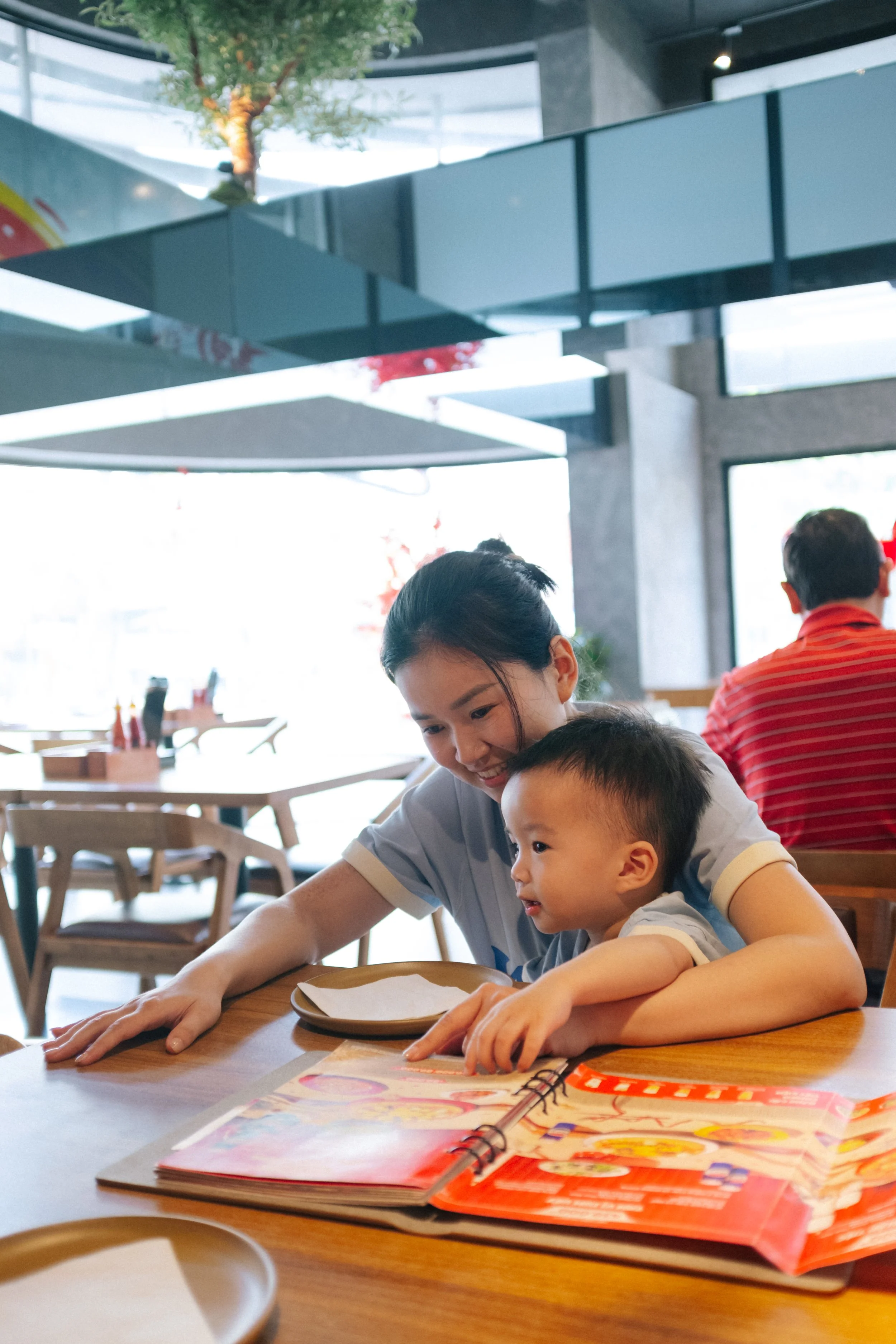 A woman and a young boy are sitting at a restaurant table, looking at a menu and smiling. The woman has dark hair tied back, and the boy is leaning on the table. The restaurant has large windows and wooden chairs.