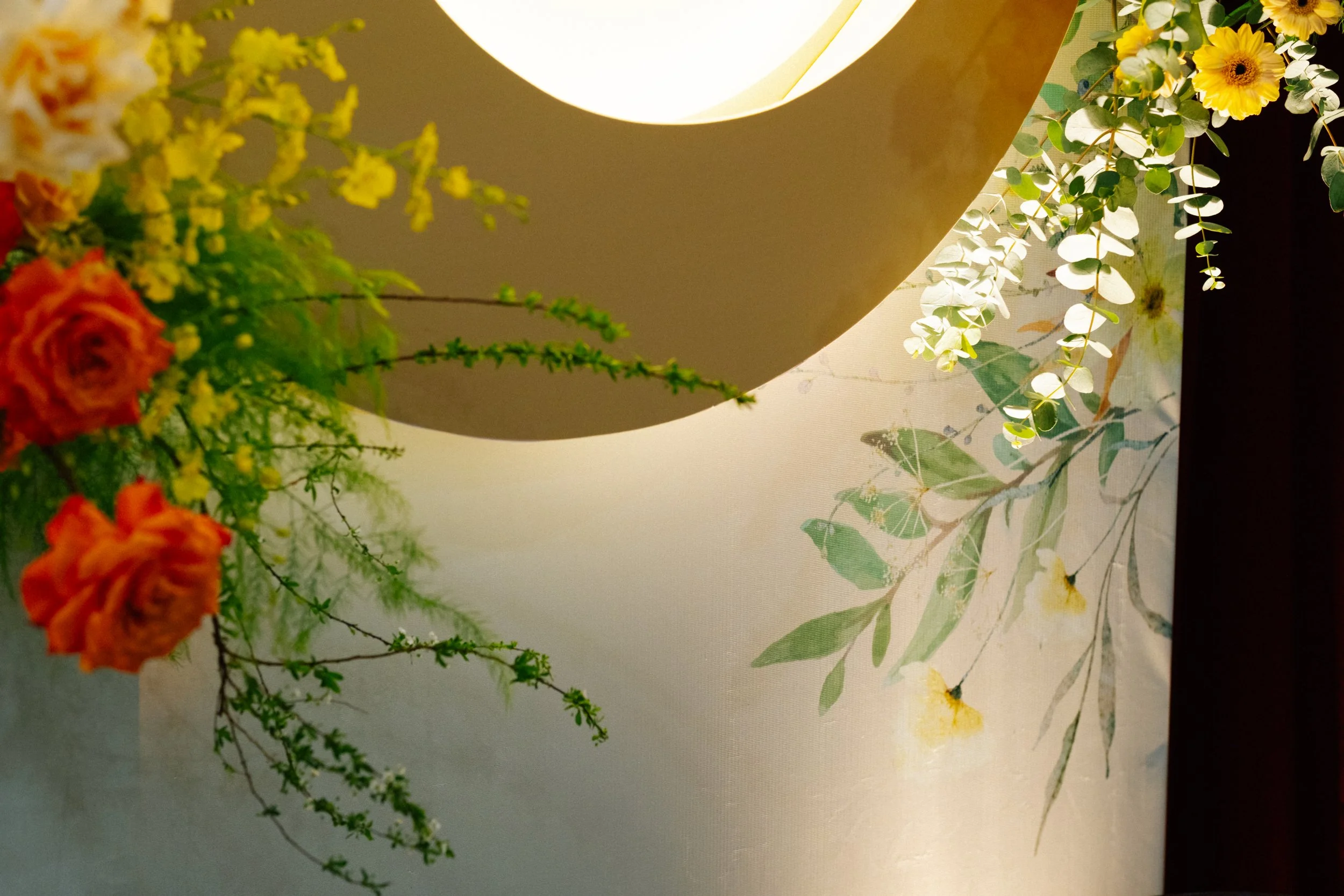 Close-up of a floral arrangement with orange roses, yellow flowers, and green foliage, decorated on a table near a large round lamp with a warm light.