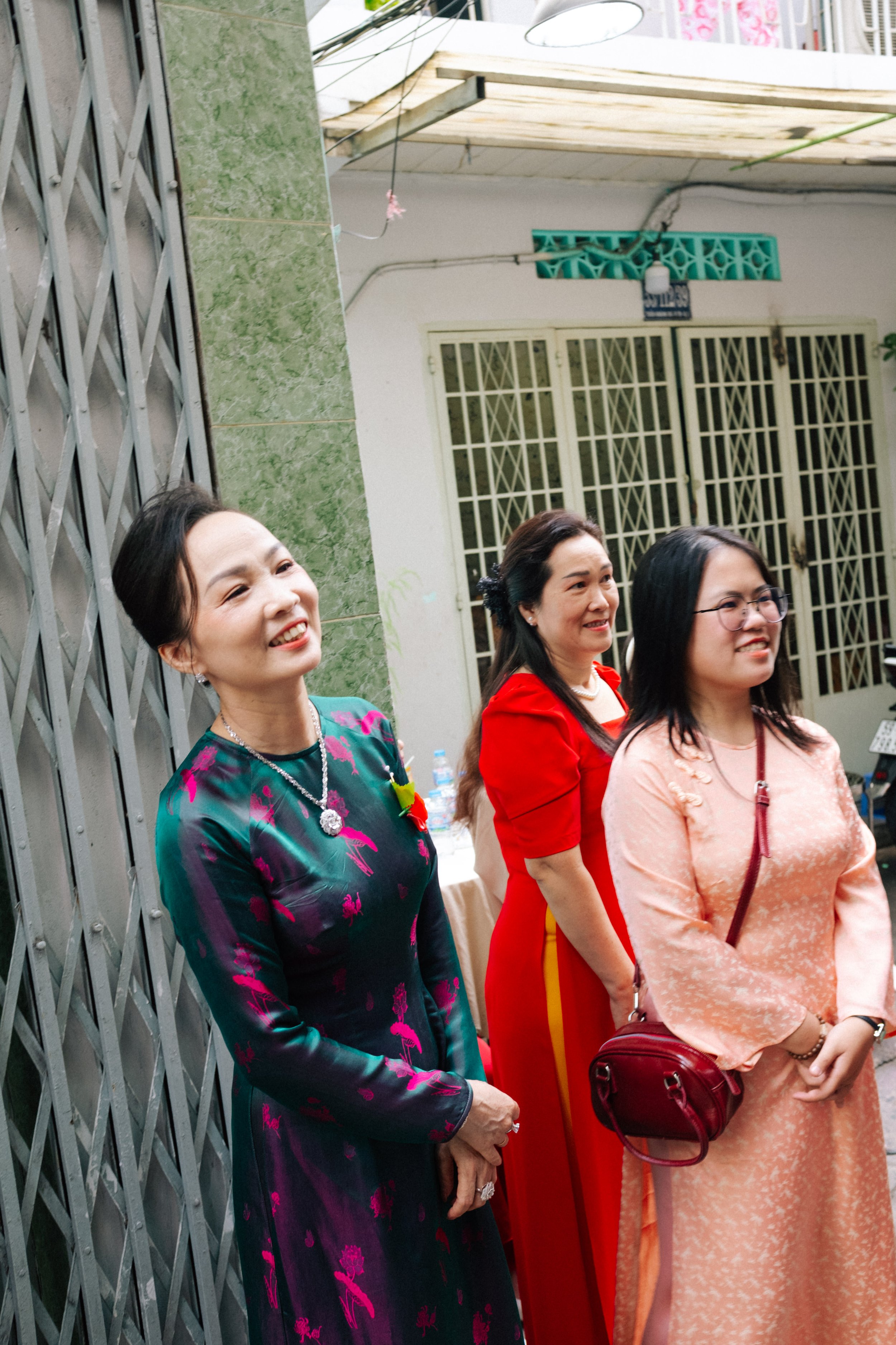 Three women in traditional attire standing outside a building with metal gates and green tiled wall.