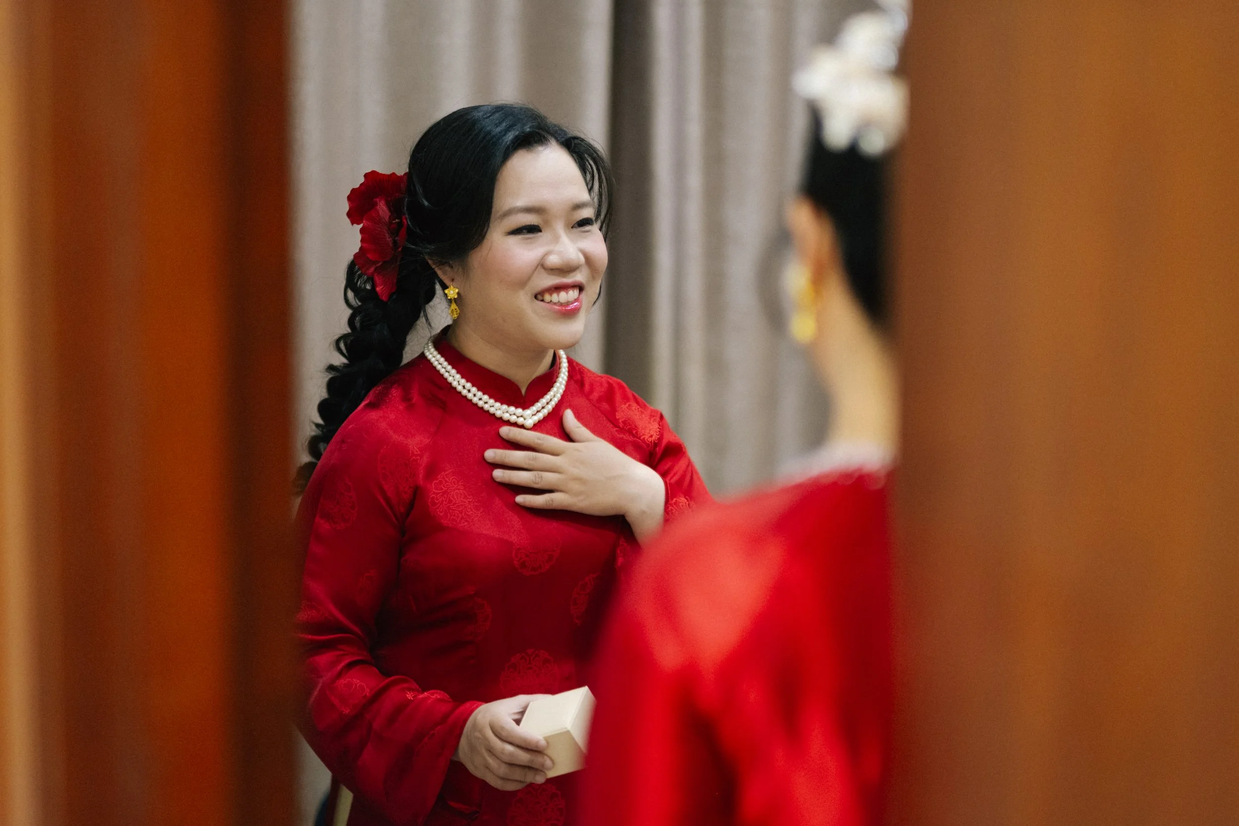 A woman in a red traditional dress, smiling and touching her chest, holding a small box, with another woman in traditional attire partially visible in the foreground.
