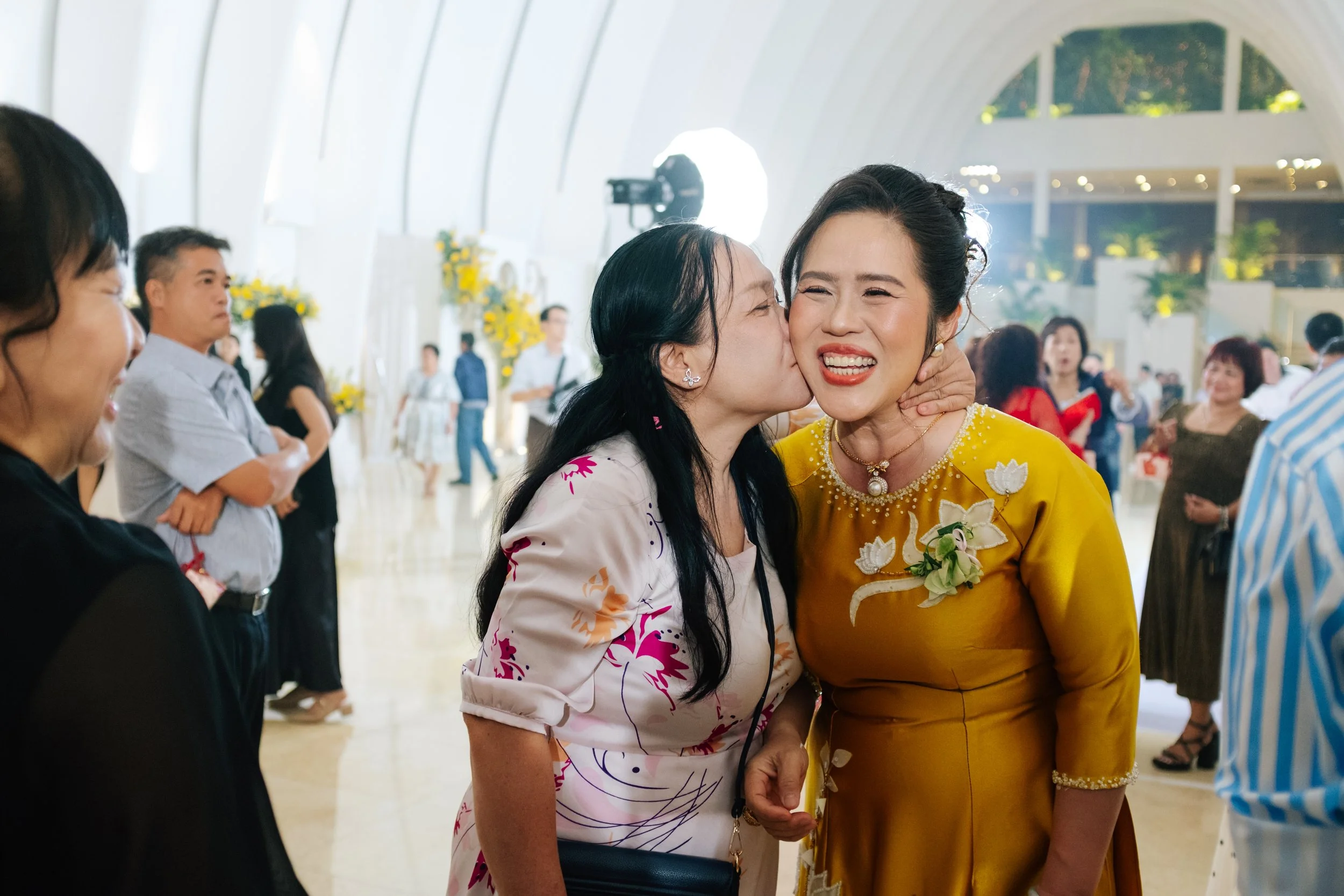 A woman in a yellow dress is being kissed on the cheek by another woman in a floral dress at a social event in a bright, modern venue with other people in the background.