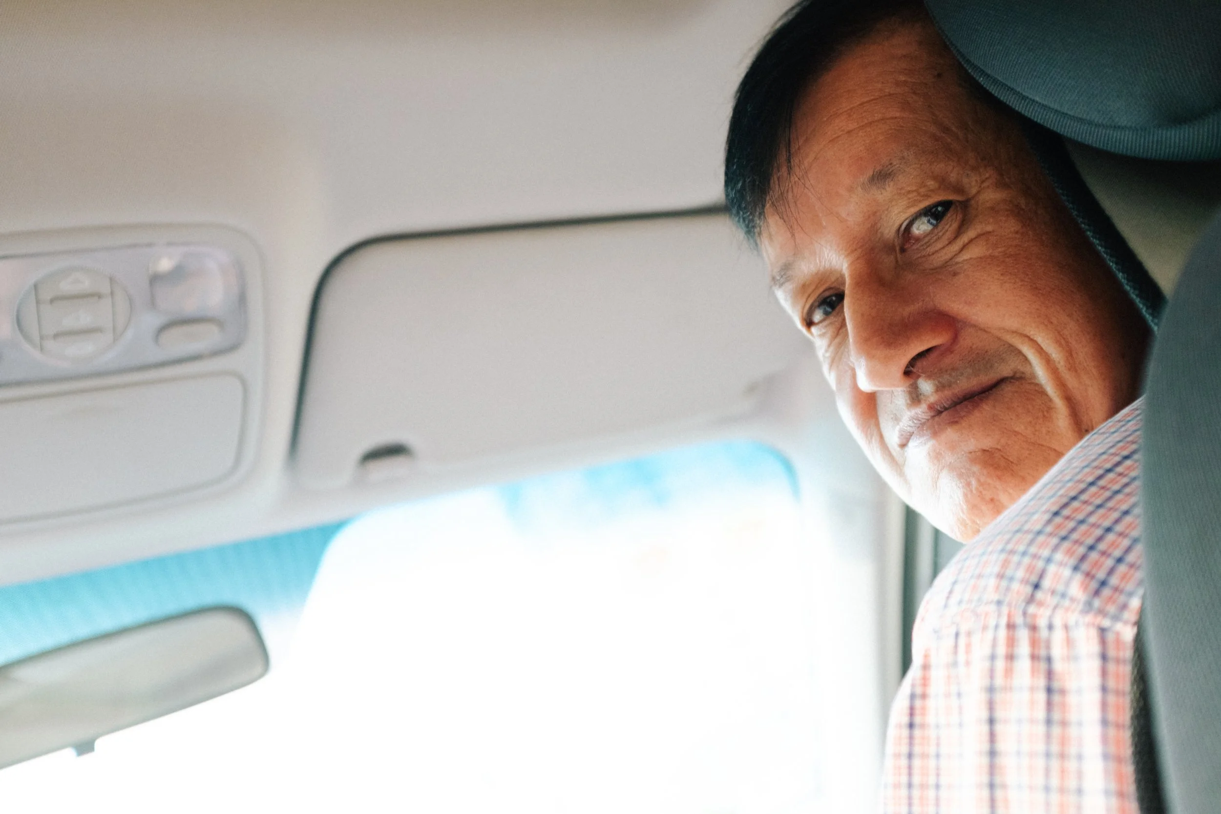Close-up of smiling middle-aged Asian man in a plaid shirt seated in a car, looking back over his shoulder.