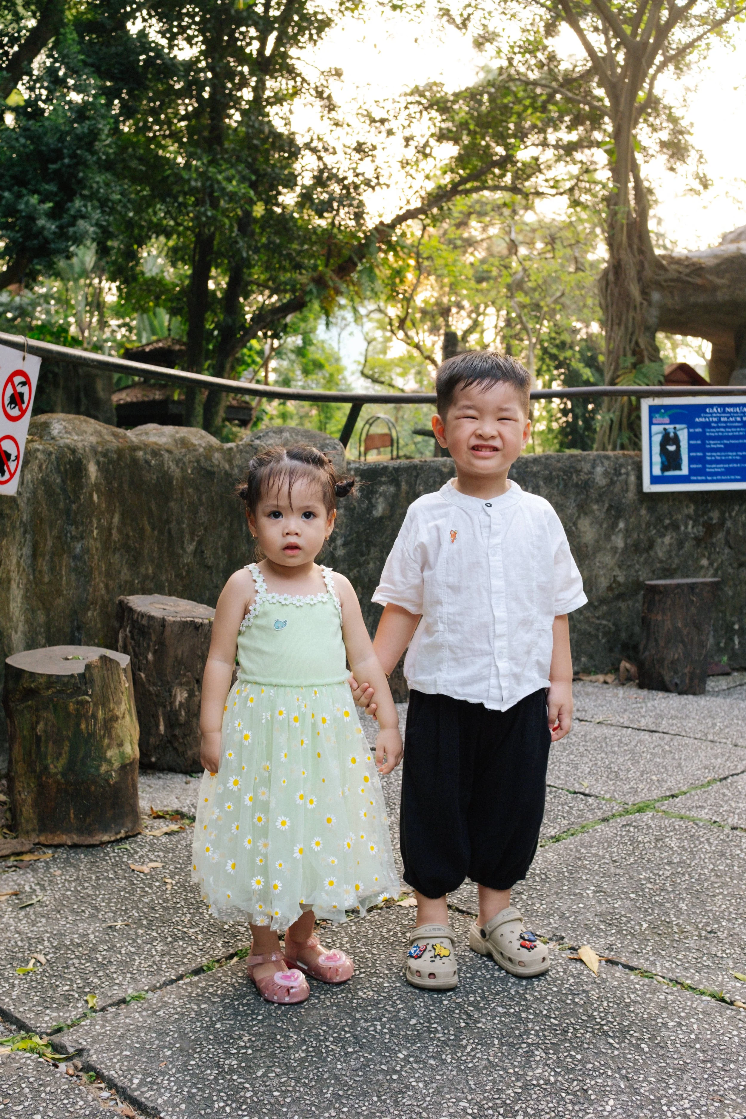 Two young children, a girl and a boy, standing outdoors on a paved area, holding hands. The girl is wearing a light green dress with white daisies and pink shoes, while the boy is dressed in a white shirt, black pants, and beige Crocs. They are in a 