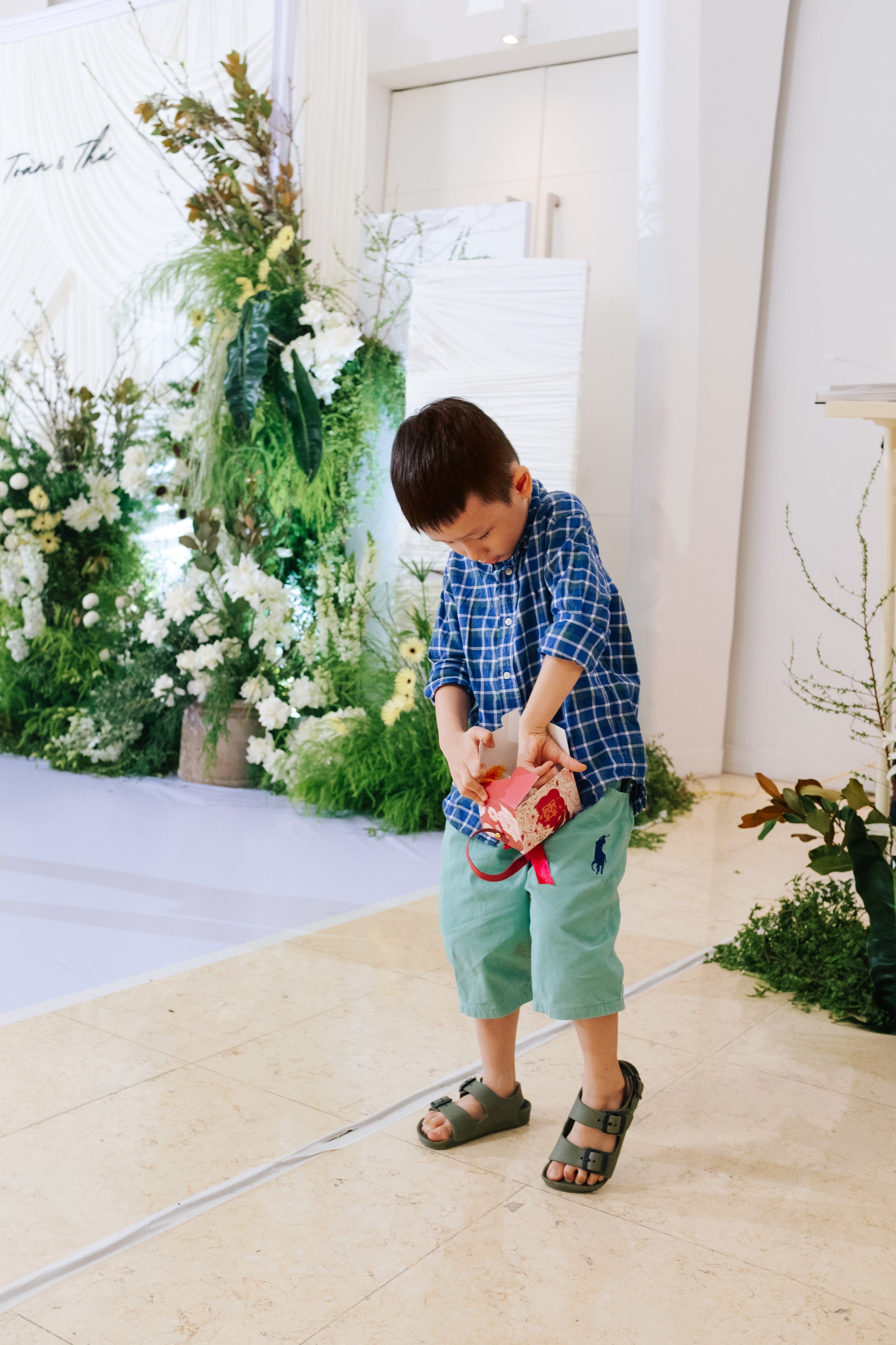 A young boy wearing a blue plaid shirt, mint green shorts, and sandals, holding a small gift box with a pink ribbon, standing inside a decorated venue with floral arrangements.