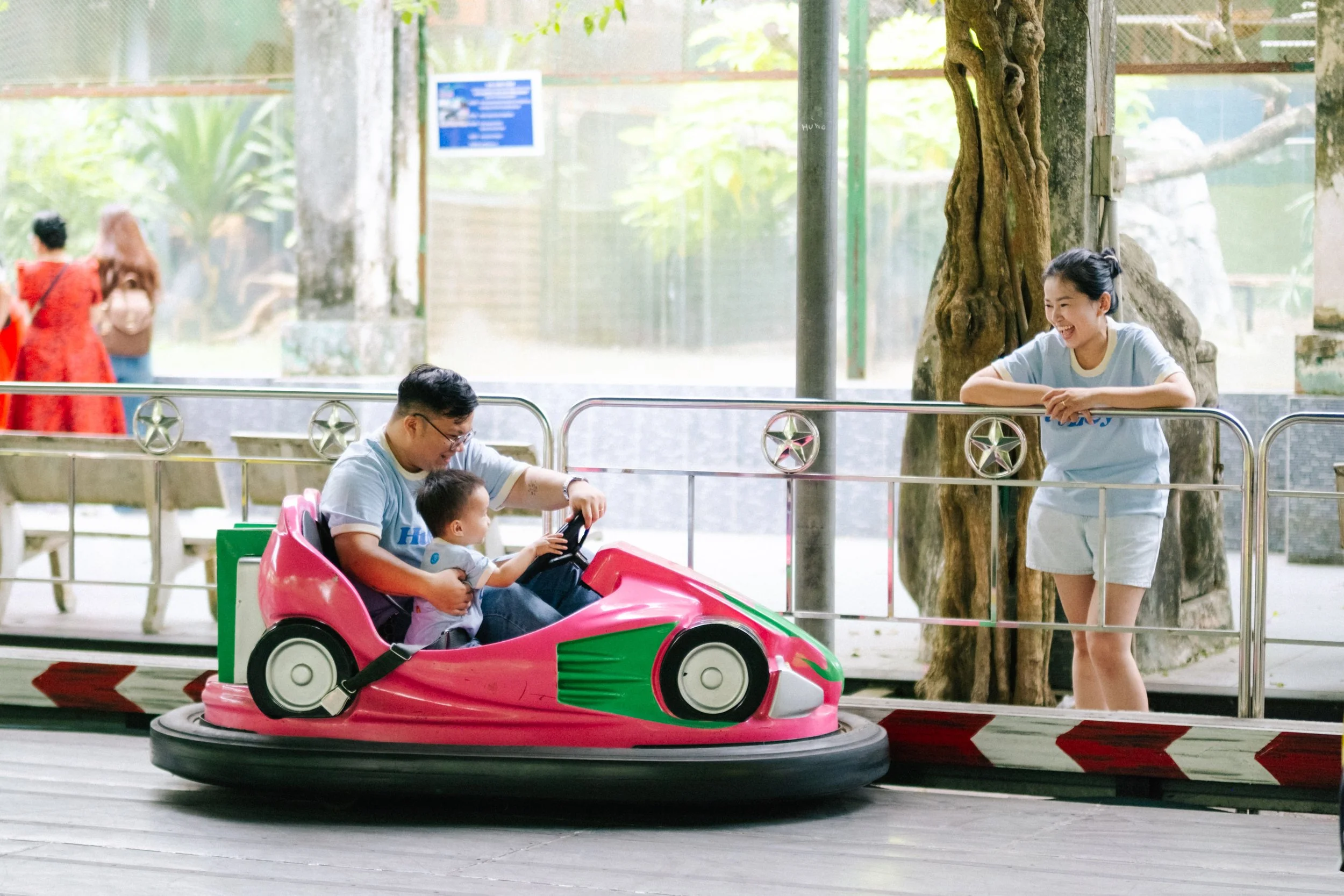 A family at an amusement park riding a pink and green bumper car, with a young woman standing nearby smiling. The scene takes place indoors with a glass window in the background.