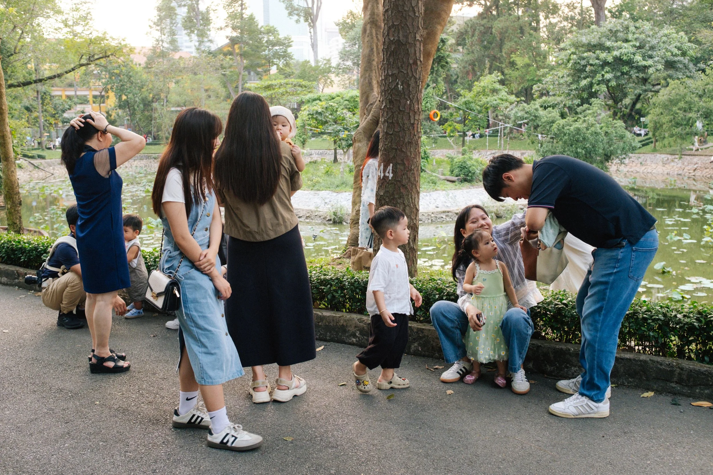 Children and adults gathering by a pond in a park, talking, and taking photos, with trees and green foliage in the background.
