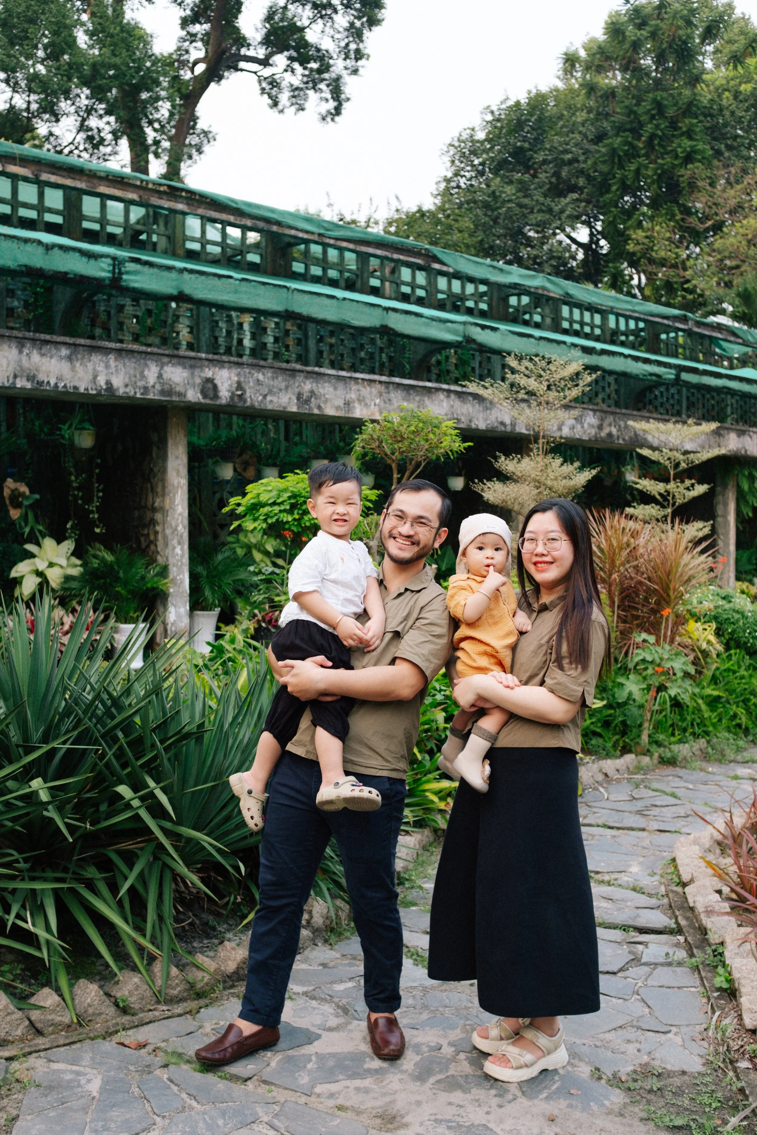 A family of four standing outdoors on a stone pathway, smiling, with lush green plants and trees in the background.