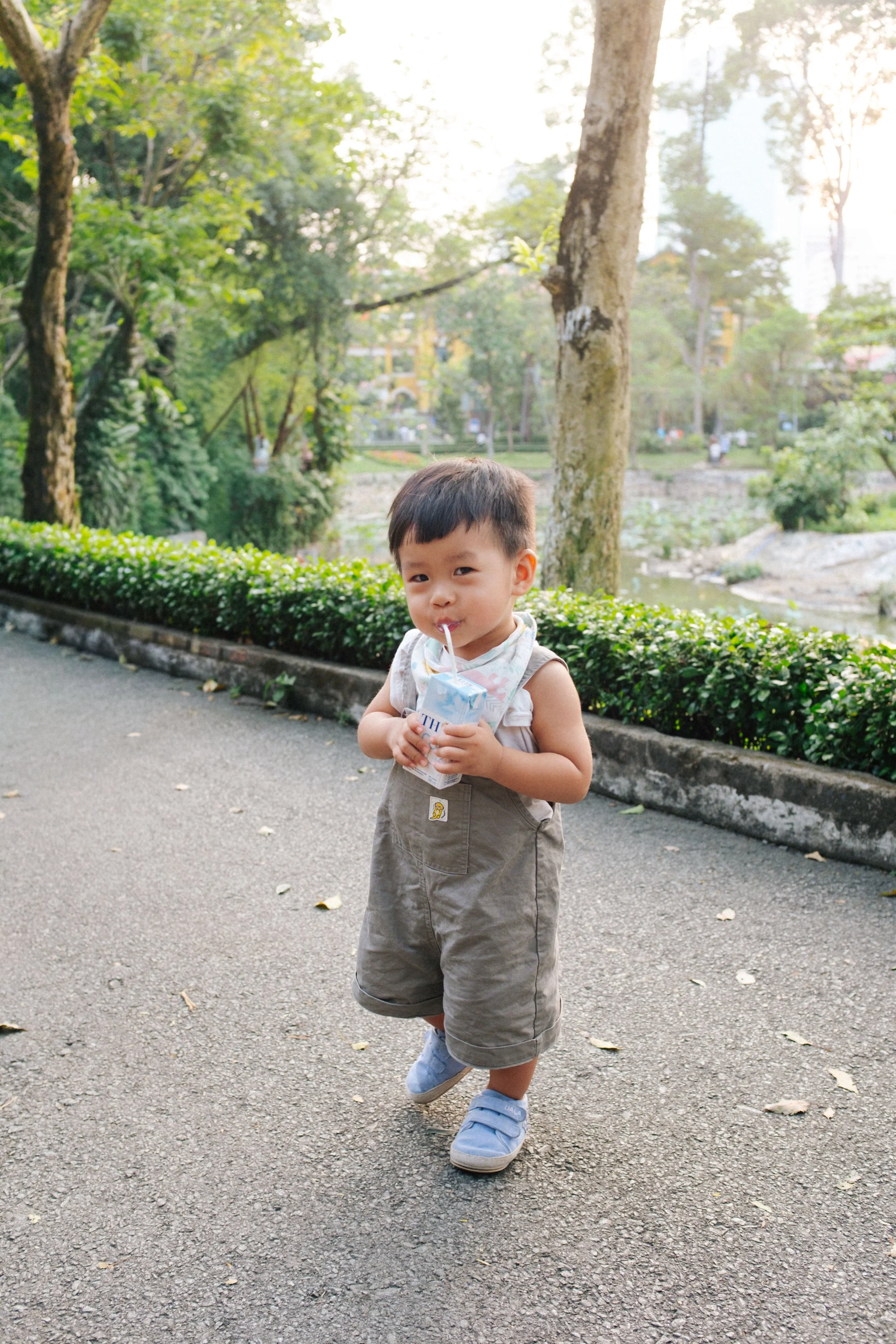 Young child standing on a paved path, holding a juice box, surrounded by trees and greenery in a park during daytime.