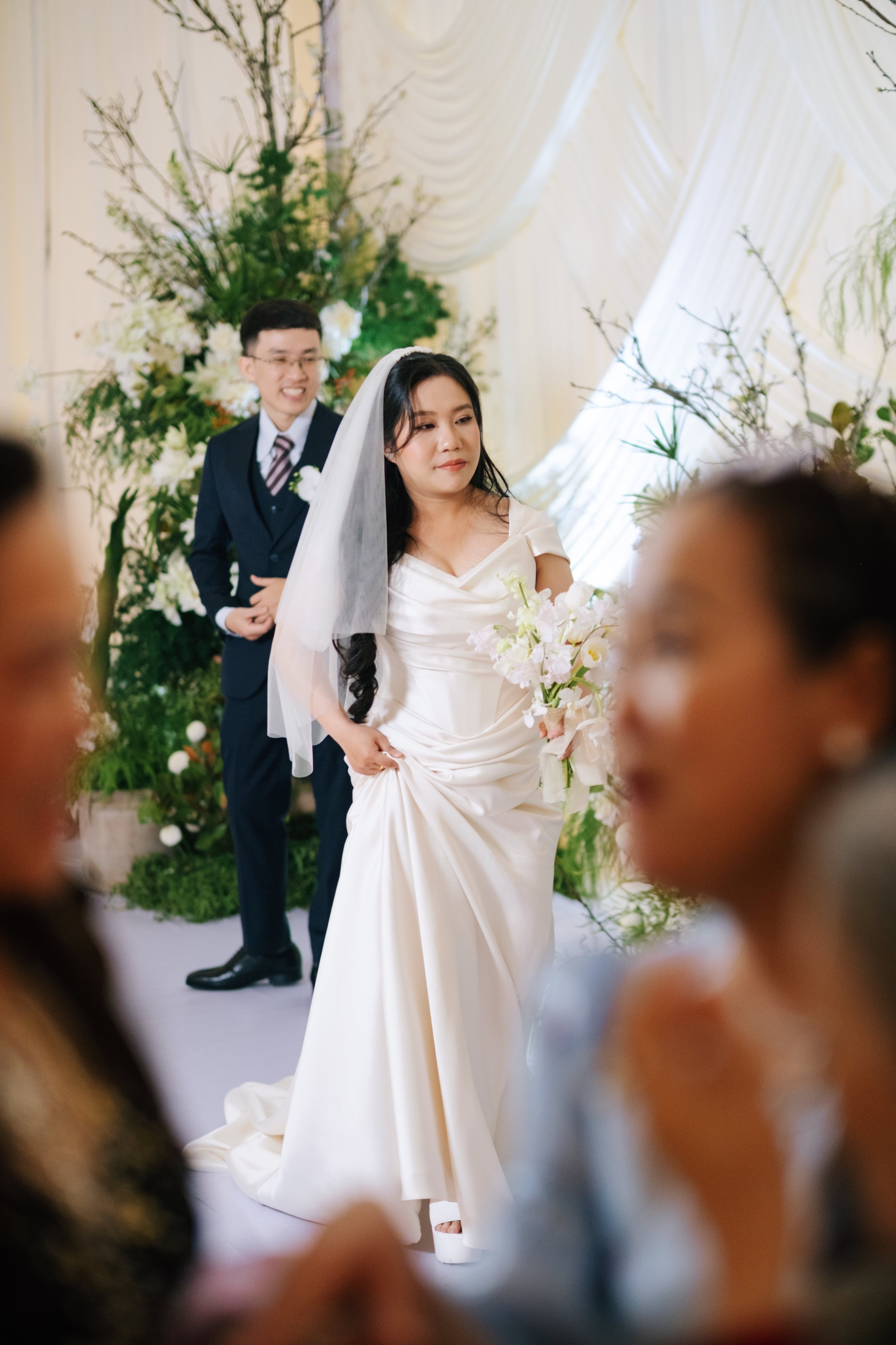 A bride in a white wedding gown holding a bouquet and a groom in a dark suit with a striped tie standing behind her in a decorated wedding venue with floral arrangements and draped fabric.