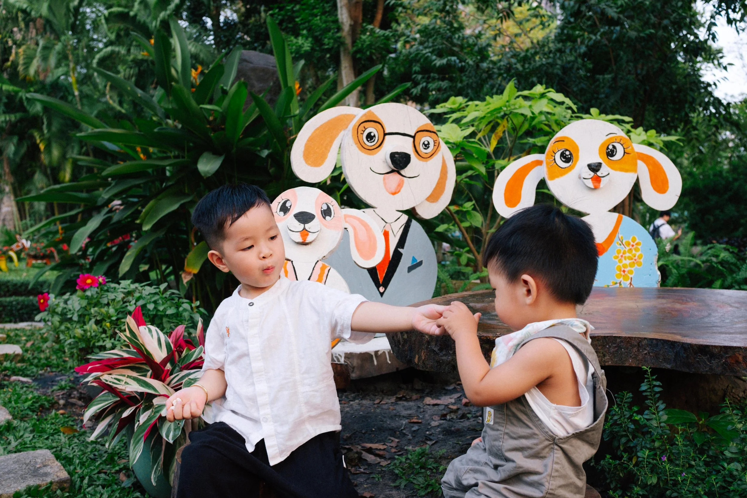 Two young boys sitting outdoors near colorful cartoon dog and cat cutouts with glasses, in a garden with green plants.