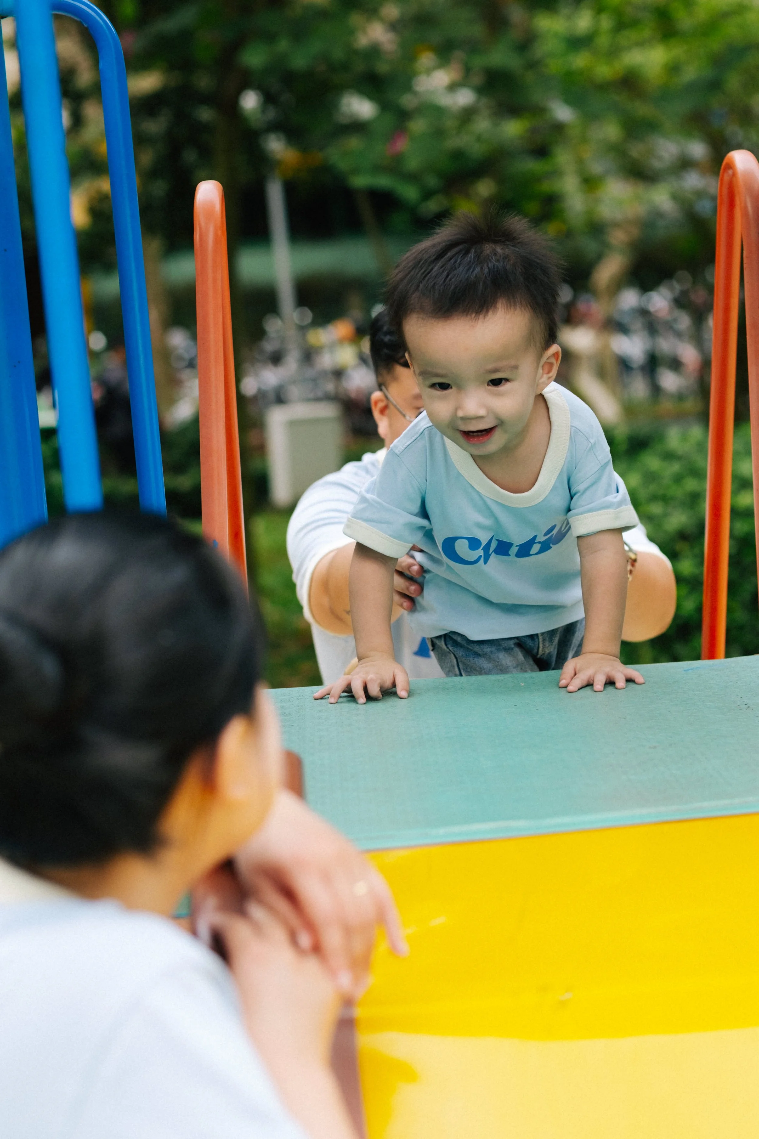 Young boy climbing over a green and yellow playground structure, smiling and looking at a woman with black hair who is sitting nearby, in a park with trees in the background.