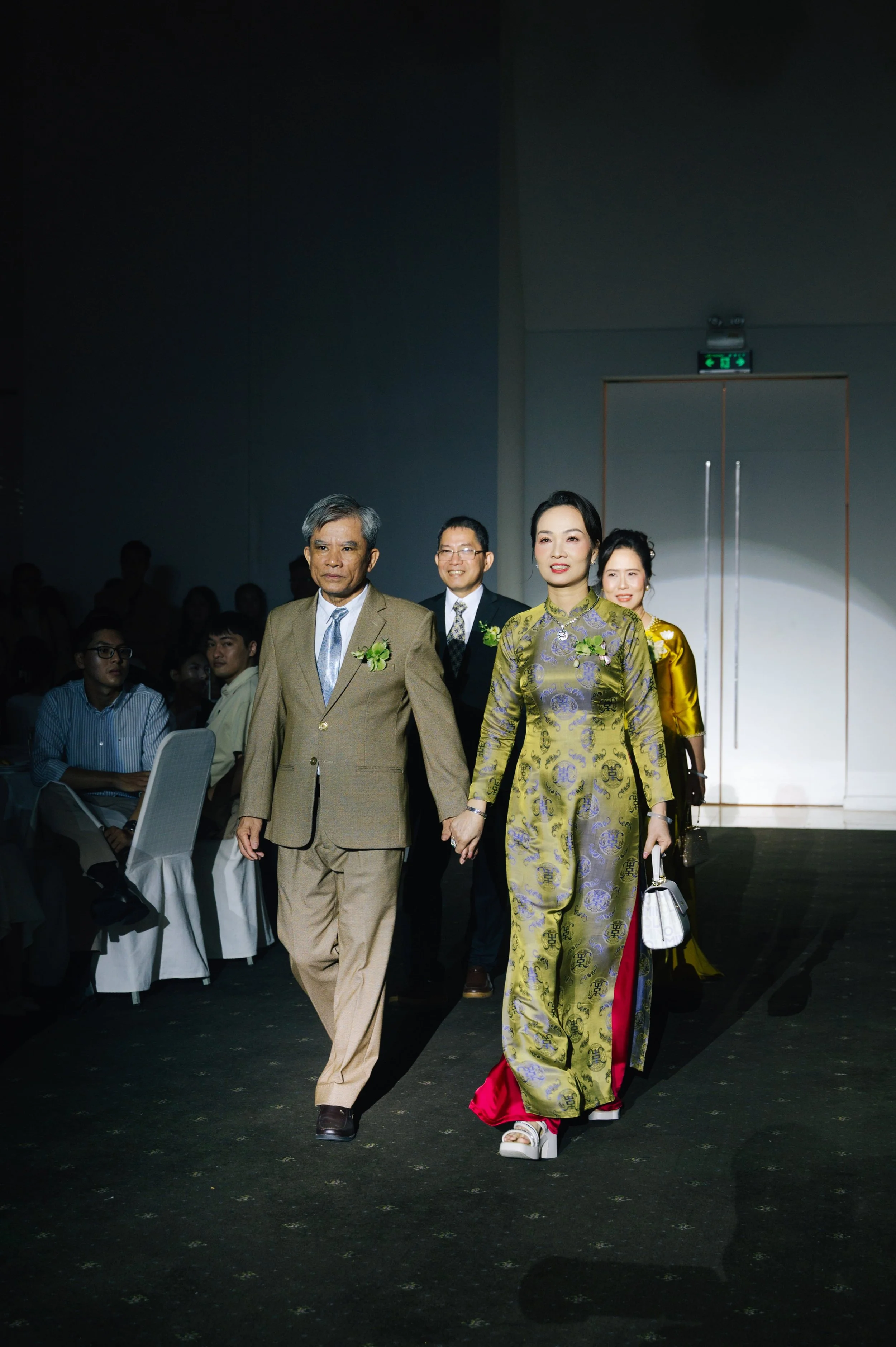 A group of people, including a woman in a traditional Asian dress, walking on a runway during a formal event, with the audience seated in the background.
