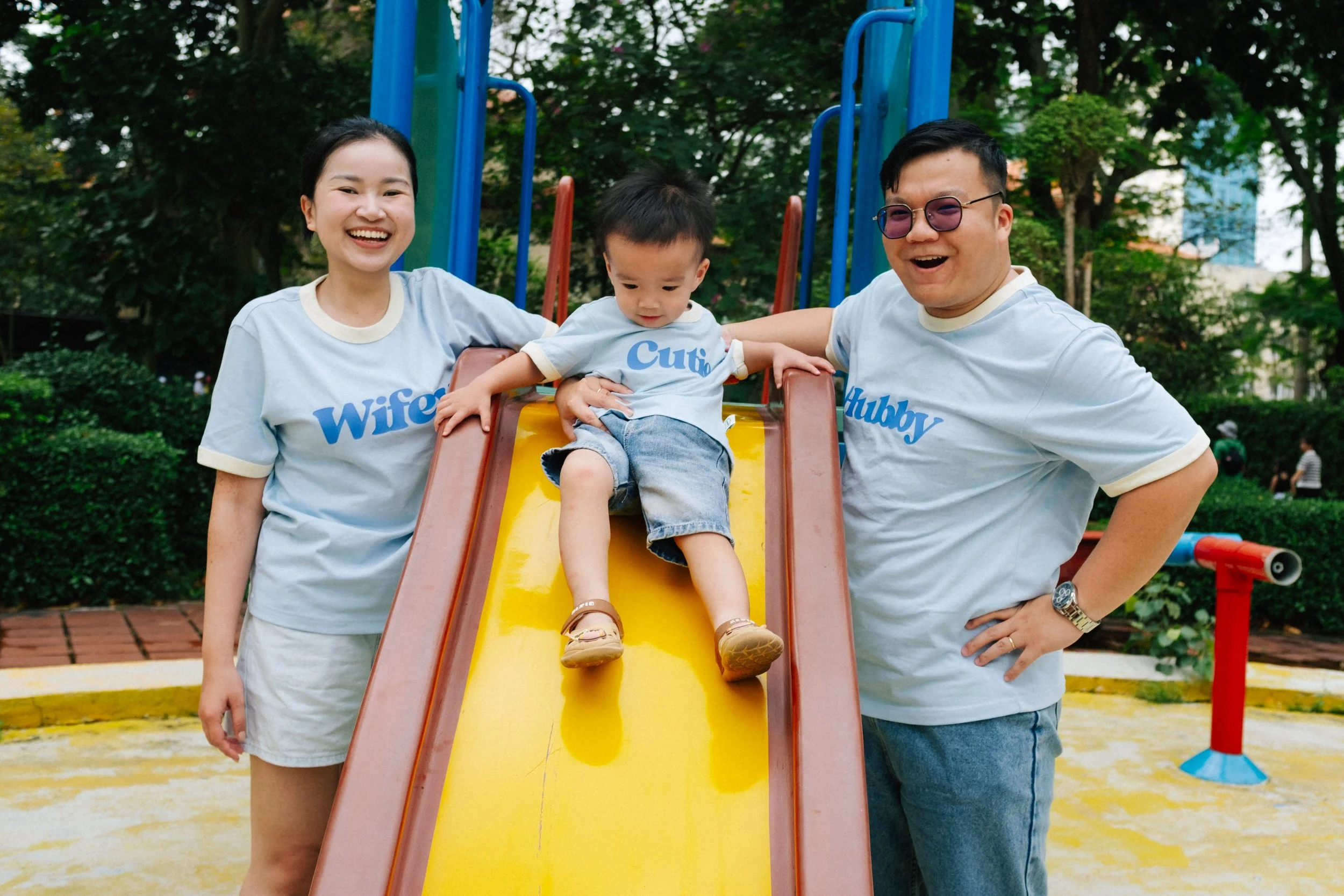 A happy family at the playground, with a woman, a man, and a young boy sitting at the top of a slide. The woman wears a shirt labeled "Wife," the boy's shirt says "Cuti," and the man's shirt says "Hubby." They are smiling and enjoying a sunny day out