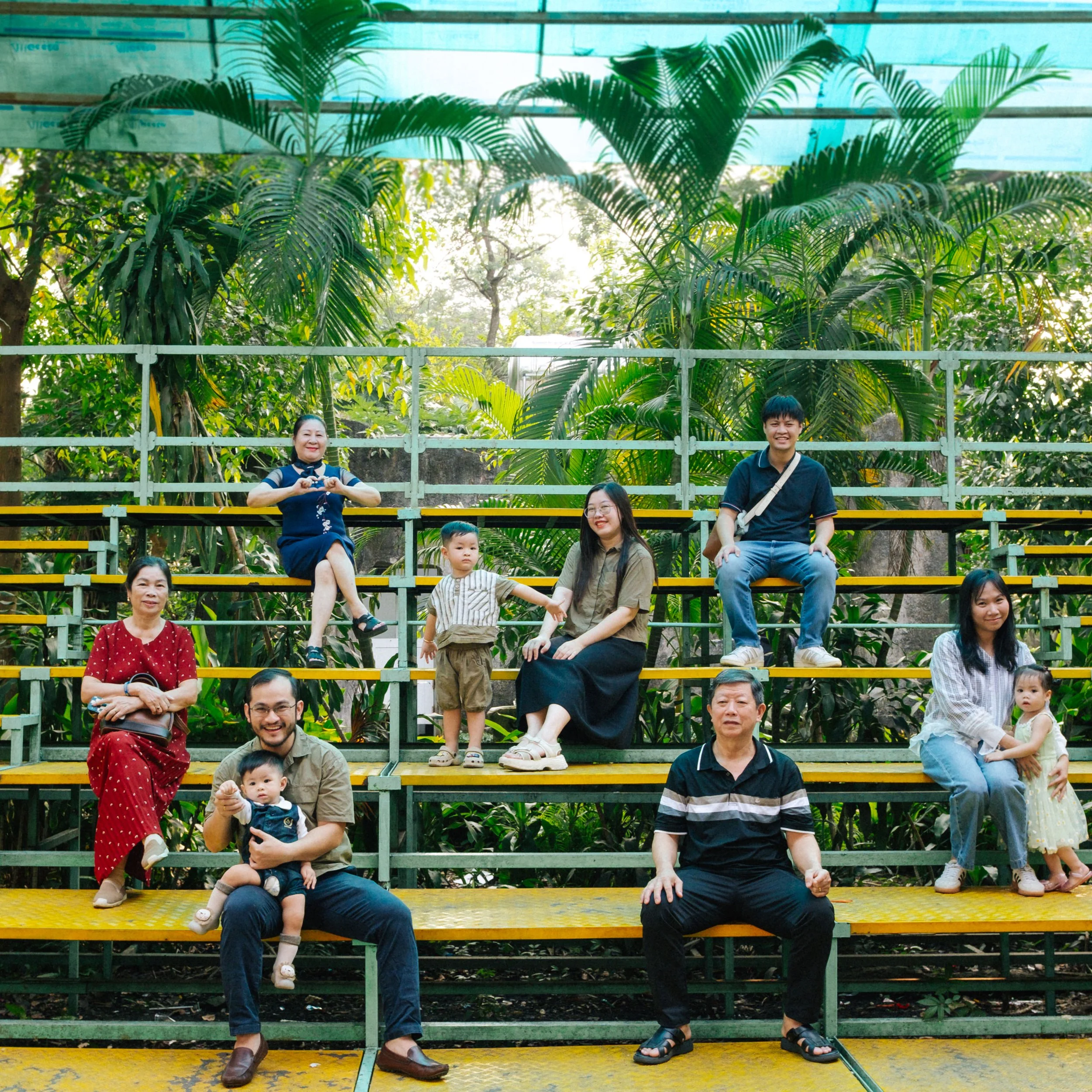 A multi-generational family group photo on outdoor bleachers with lush green tropical plants in the background.