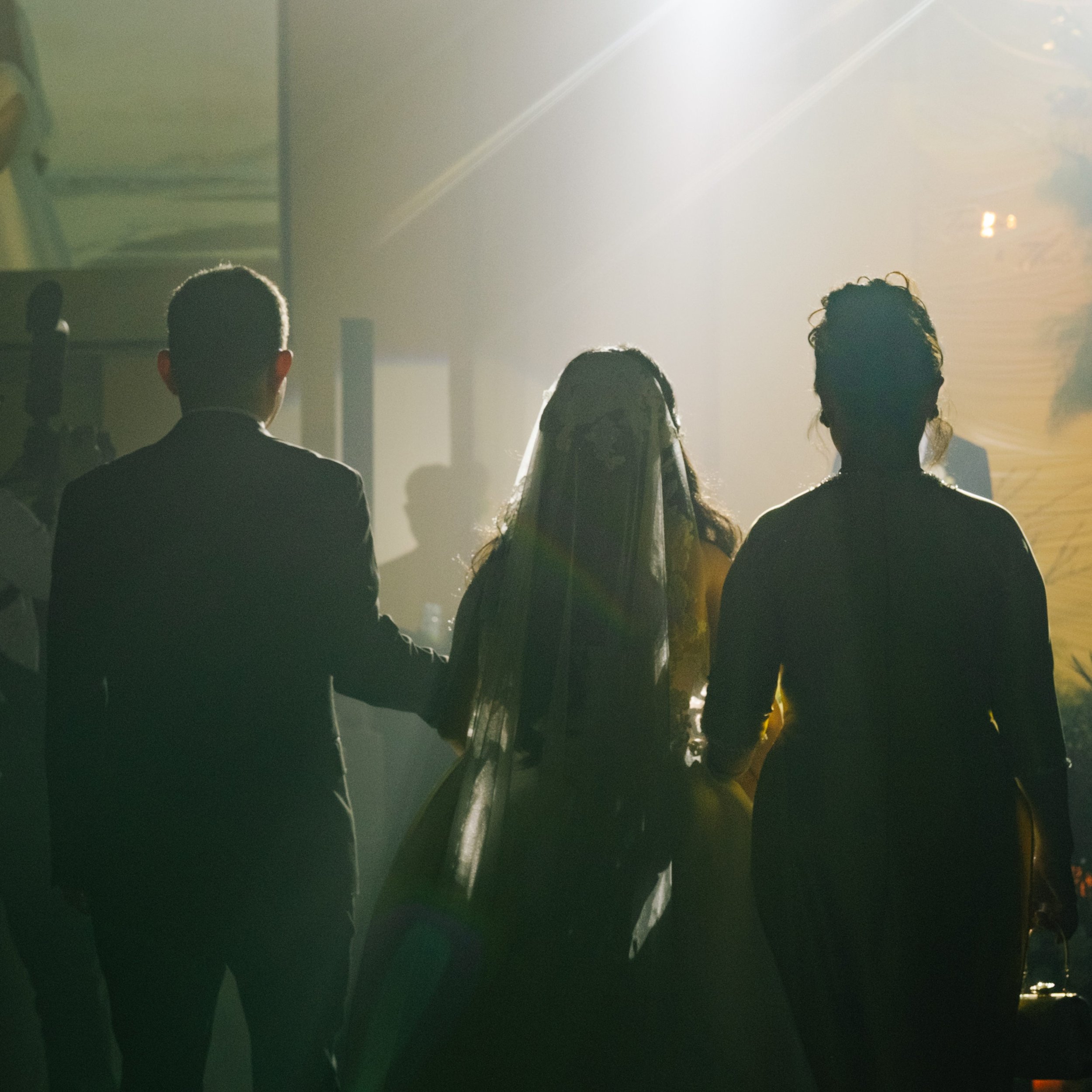 Silhouettes of three people standing together in a well-lit indoor space, including a woman in a wedding gown and veil, hinting at a wedding or celebration.