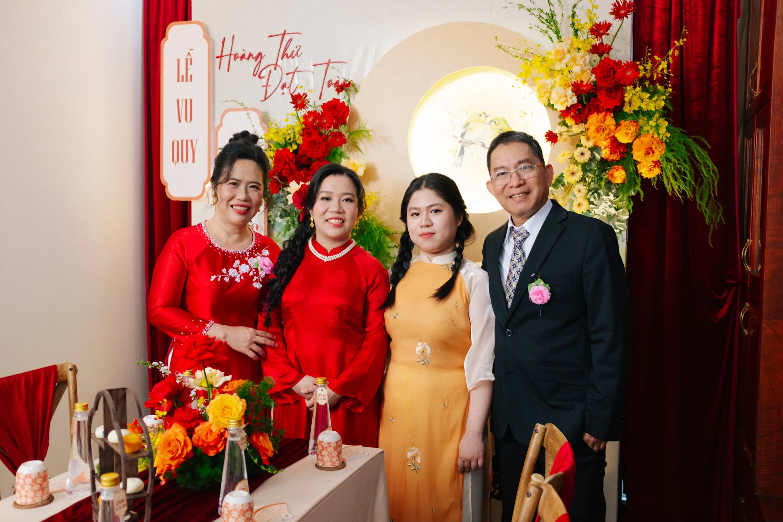 A family gathered at a traditional Vietnamese celebration, standing in front of a decorated backdrop with colorful flowers and a circular art piece, wearing traditional and formal attire.