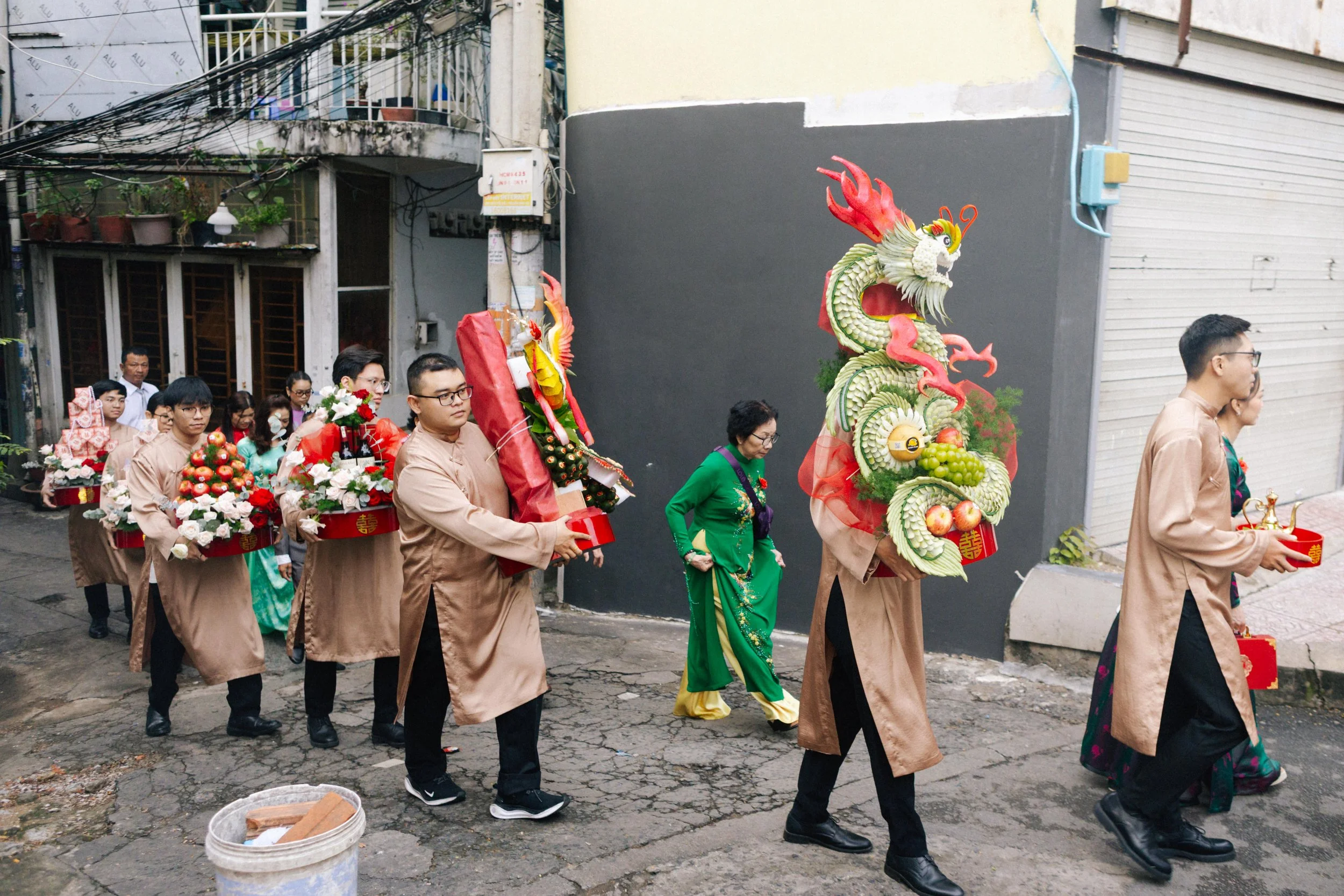 People participating in a cultural procession, carrying elaborate dragon and fruit-themed decorations, on a street in an urban area.