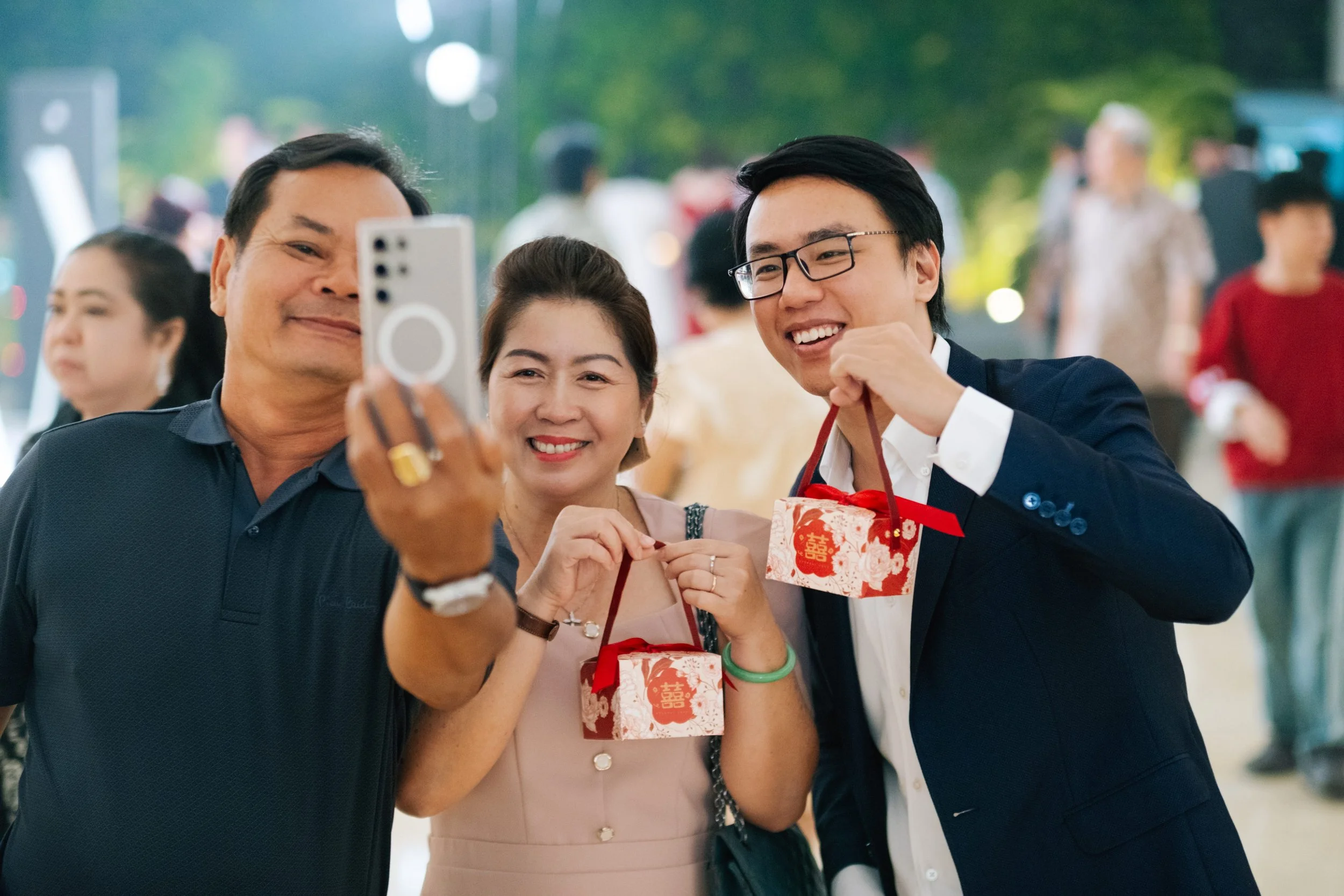 Three people taking a selfie at a celebration, each holding a gift bag with red and gold designs, smiling and dressed nicely, with a blurred crowd in the background.