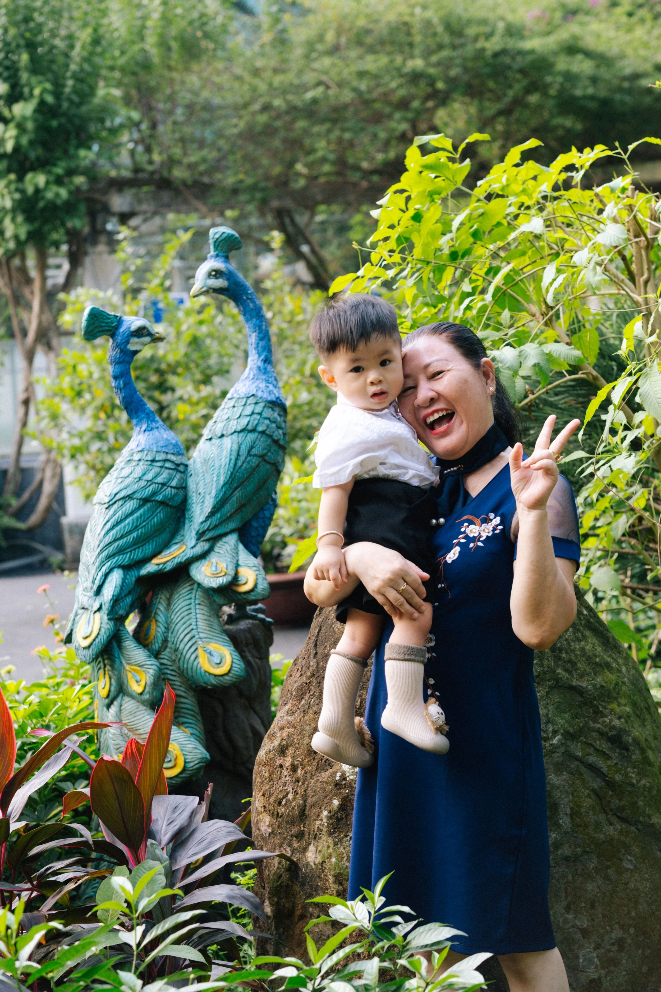 A woman and a young boy smiling and making a peace sign while standing outdoors near a rock and a colorful peacock sculpture, surrounded by lush green plants and trees.