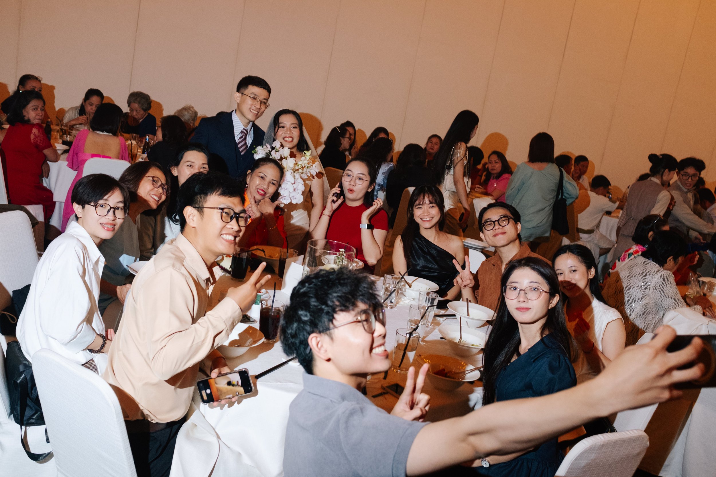 Group of people at a celebration or wedding banquet, smiling, taking selfies, and enjoying the event in a large event hall.