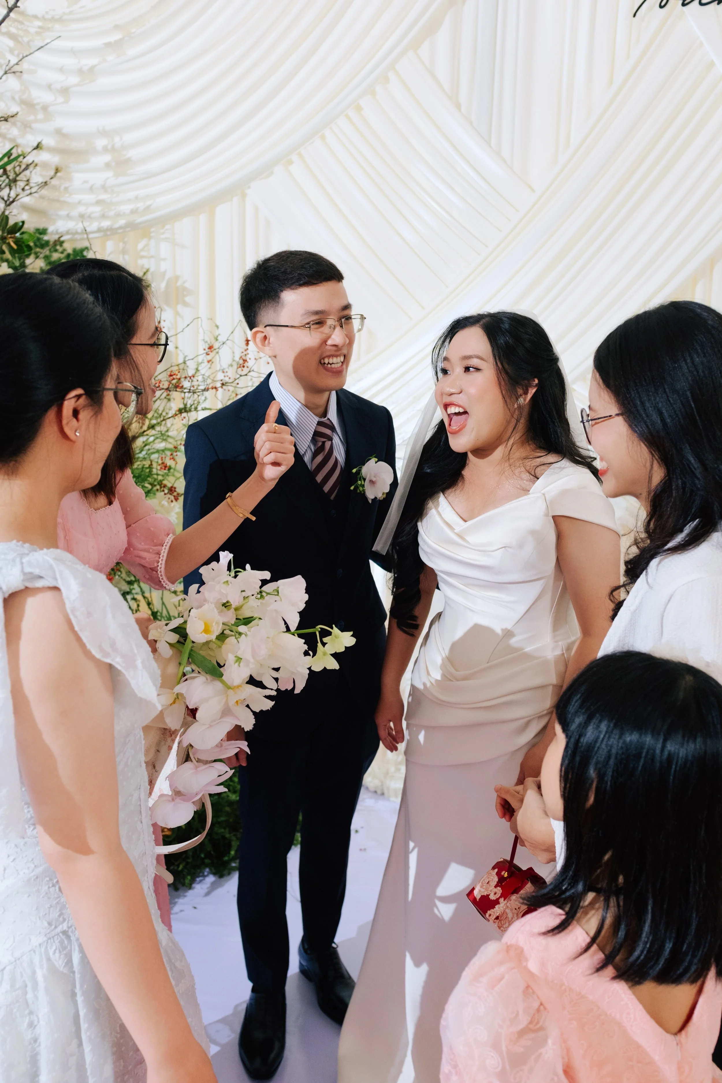 A wedding scene with a bride and groom sharing a joyful moment surrounded by friends. The bride is wearing a white dress and smiling, while the groom in a navy suit and striped tie also smiling. Friends dressed in pastel-colored dresses hold flowers 