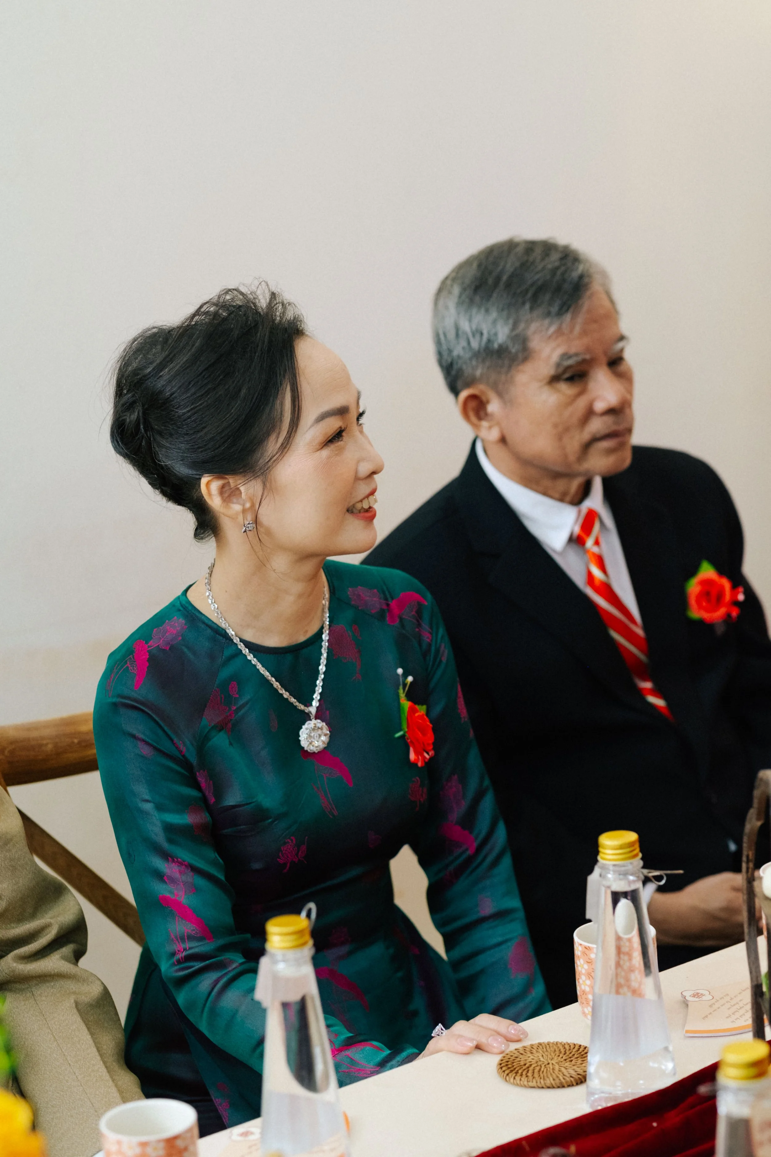 A woman in a teal dress with floral patterns, wearing a necklace and earrings, sitting beside a man in a black suit with a red and white striped tie, both with red floral lapel pins, at a formal event.