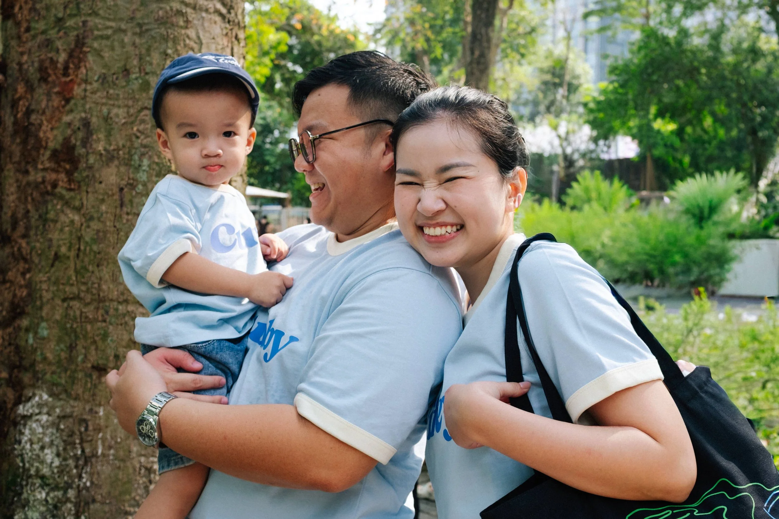 A smiling woman, a man holding a young boy, outdoor park setting with trees and greenery.