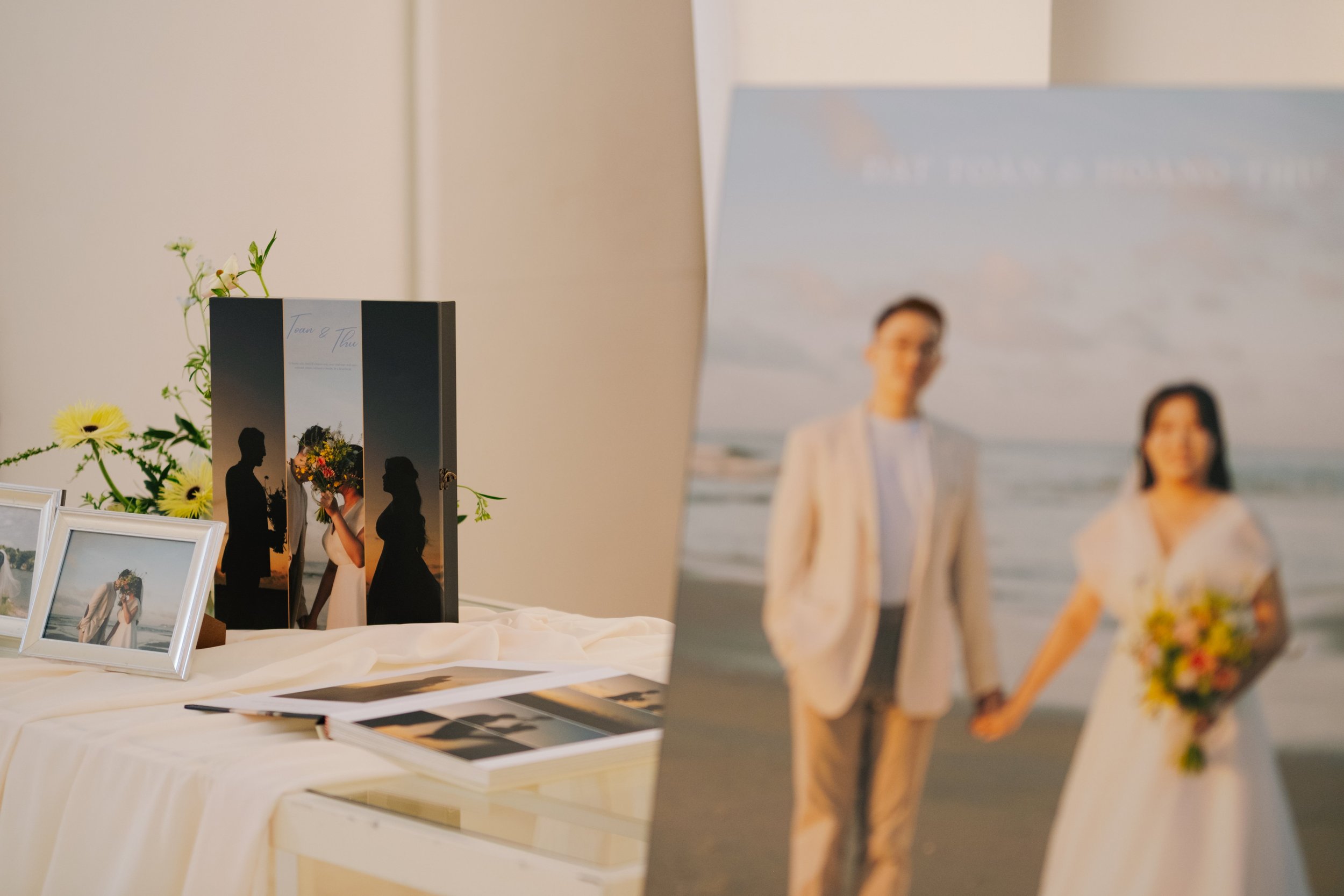 Display table with framed photographs and a silver folding screen showing a wedding photo of a couple holding a bouquet and holding hands on the beach.