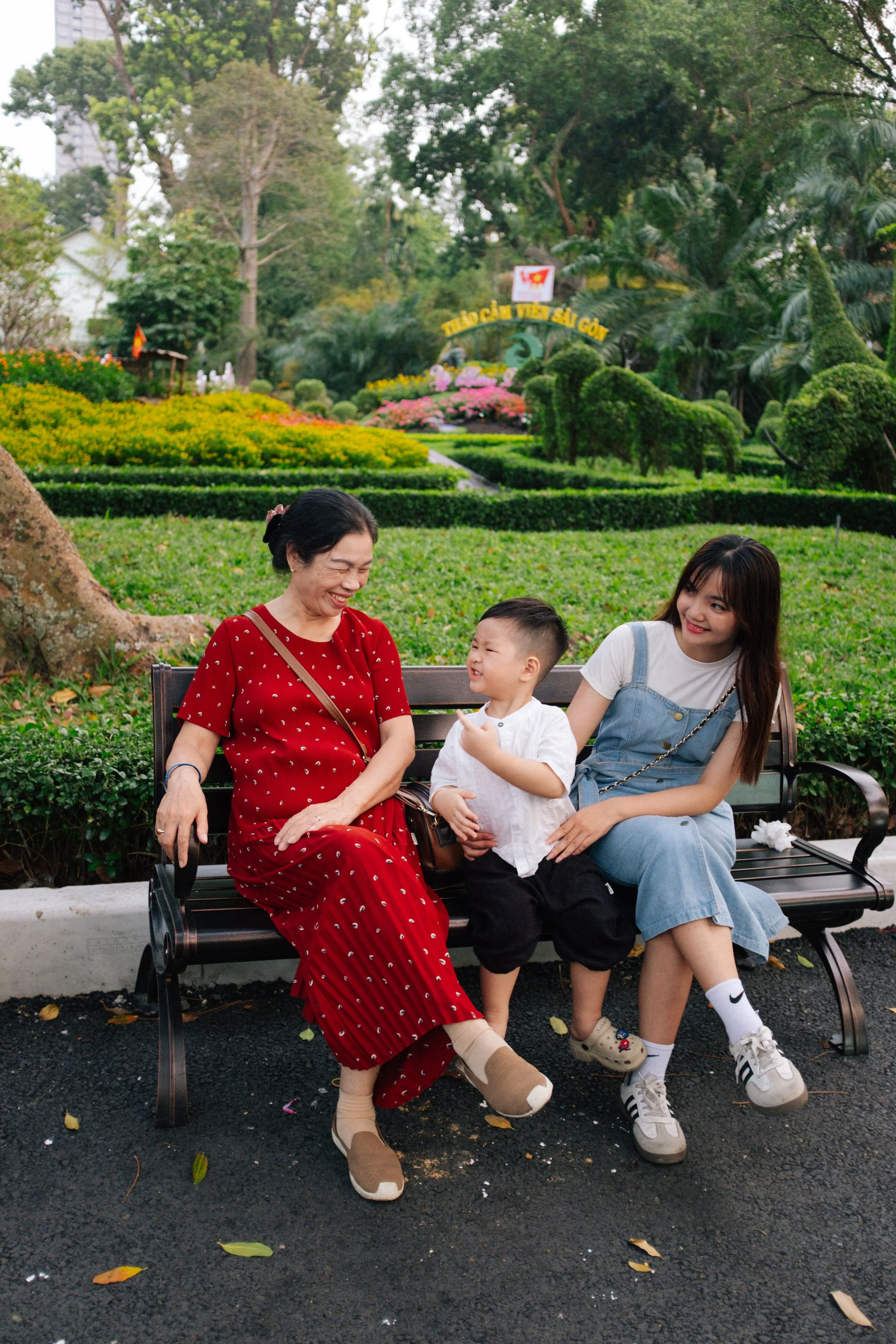 A woman, a little boy, and a young woman sitting on a park bench, laughing and smiling, with a lush garden and topiary animals in the background.