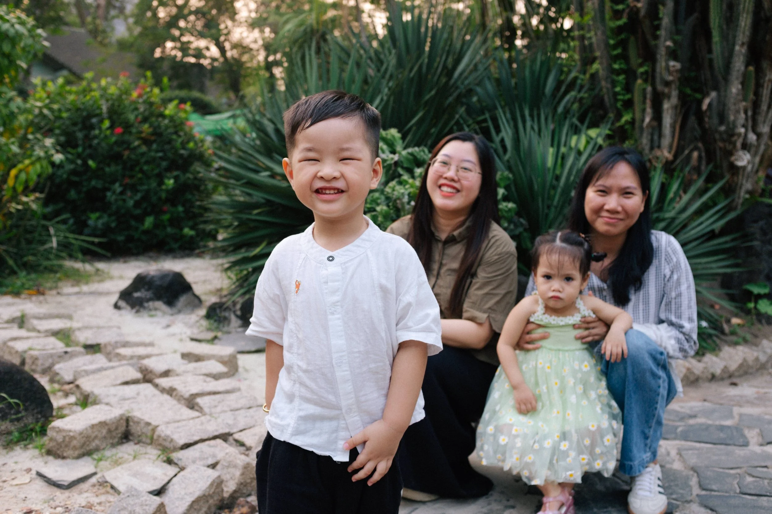 Four people, two women and two children, are sitting and standing on a stone pathway in a garden with green bushes and trees in the background. The boy in front is smiling and wearing a white shirt, while the girl in a green dress looks serious. The 