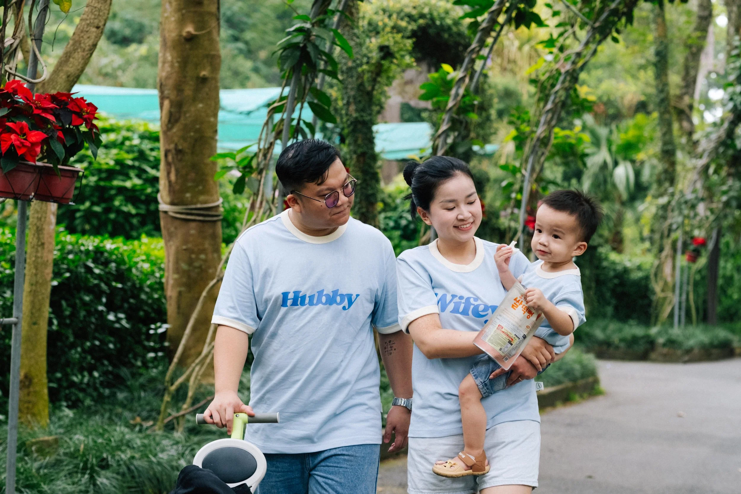 A family of three, with a man, woman, and a young child, walking in a lush, green outdoor garden or park. They are wearing matching light blue T-shirts labeled "Hubby" and "Wife." The woman is holding the child, who appears to be eating a snack and i