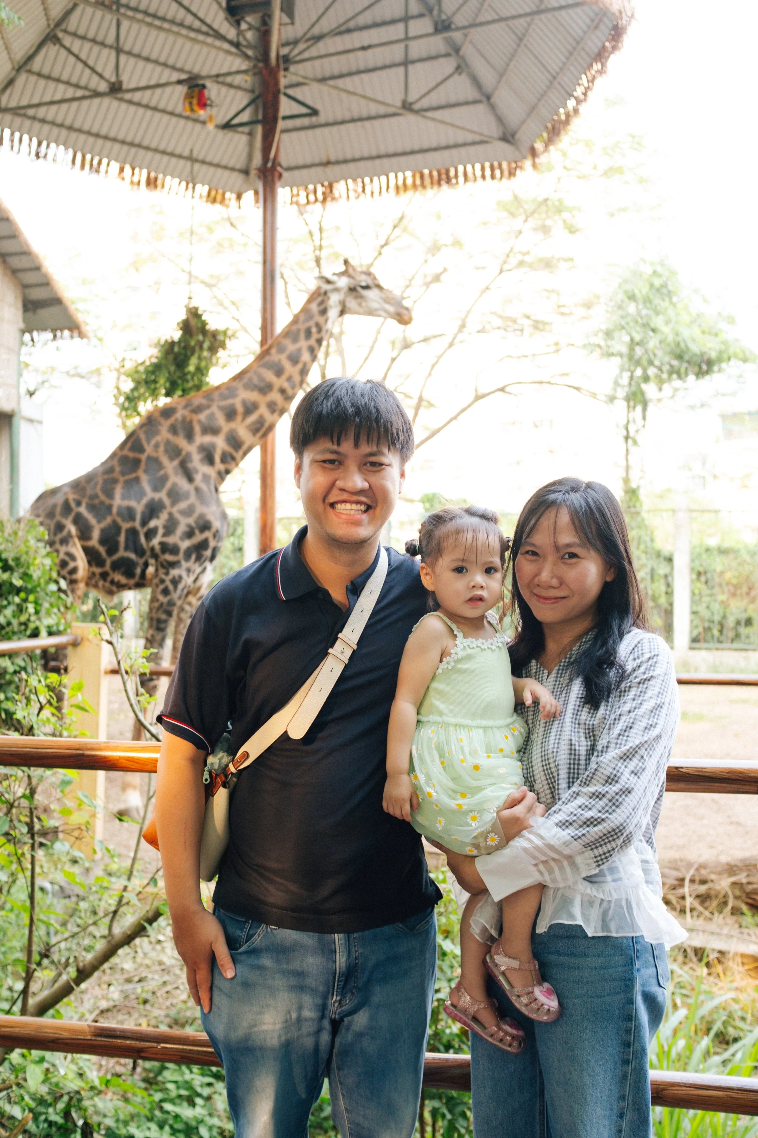 A family of three visiting a zoo, with a giraffe in the background. The father is smiling, wearing a black shirt, and standing next to a woman holding a young girl, who is looking at the camera. The three are standing near a wooden railing with green