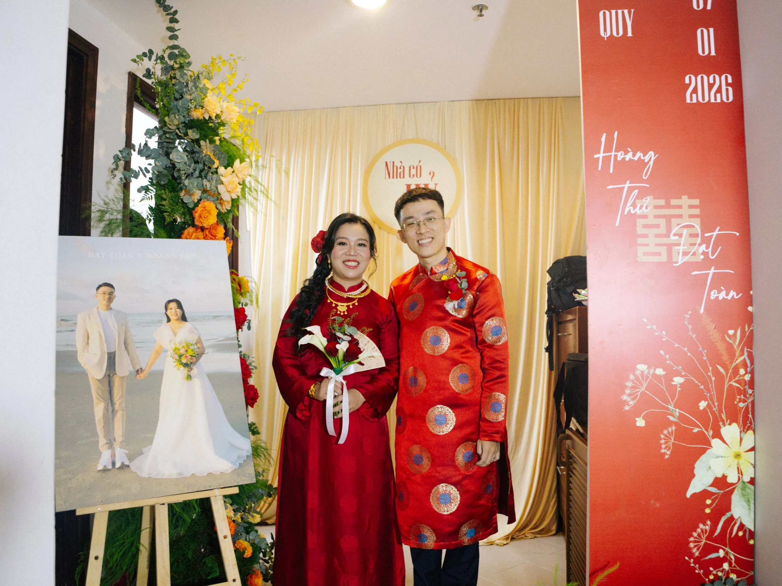 A couple dressed in traditional Vietnamese red wedding attire posing for a photo indoors, standing in front of a backdrop with yellow curtains, surrounded by floral decorations, with a wedding photo on an easel to their left and a red vertical banner