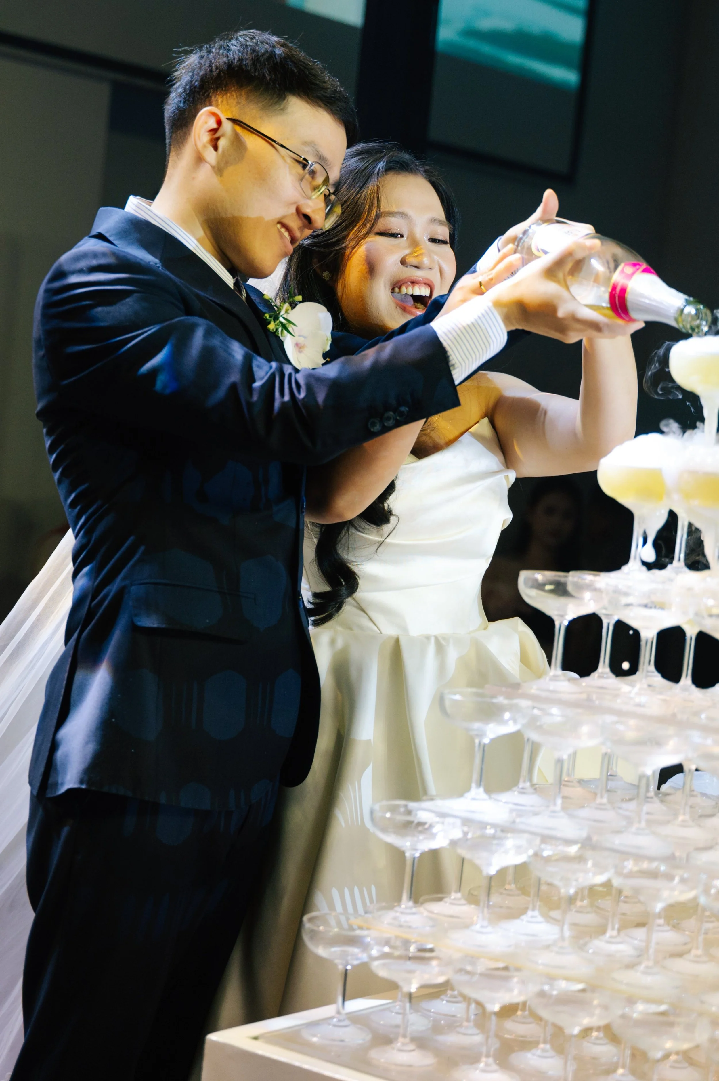 A couple in wedding attire pouring champagne into a pyramid of glasses at their wedding reception.